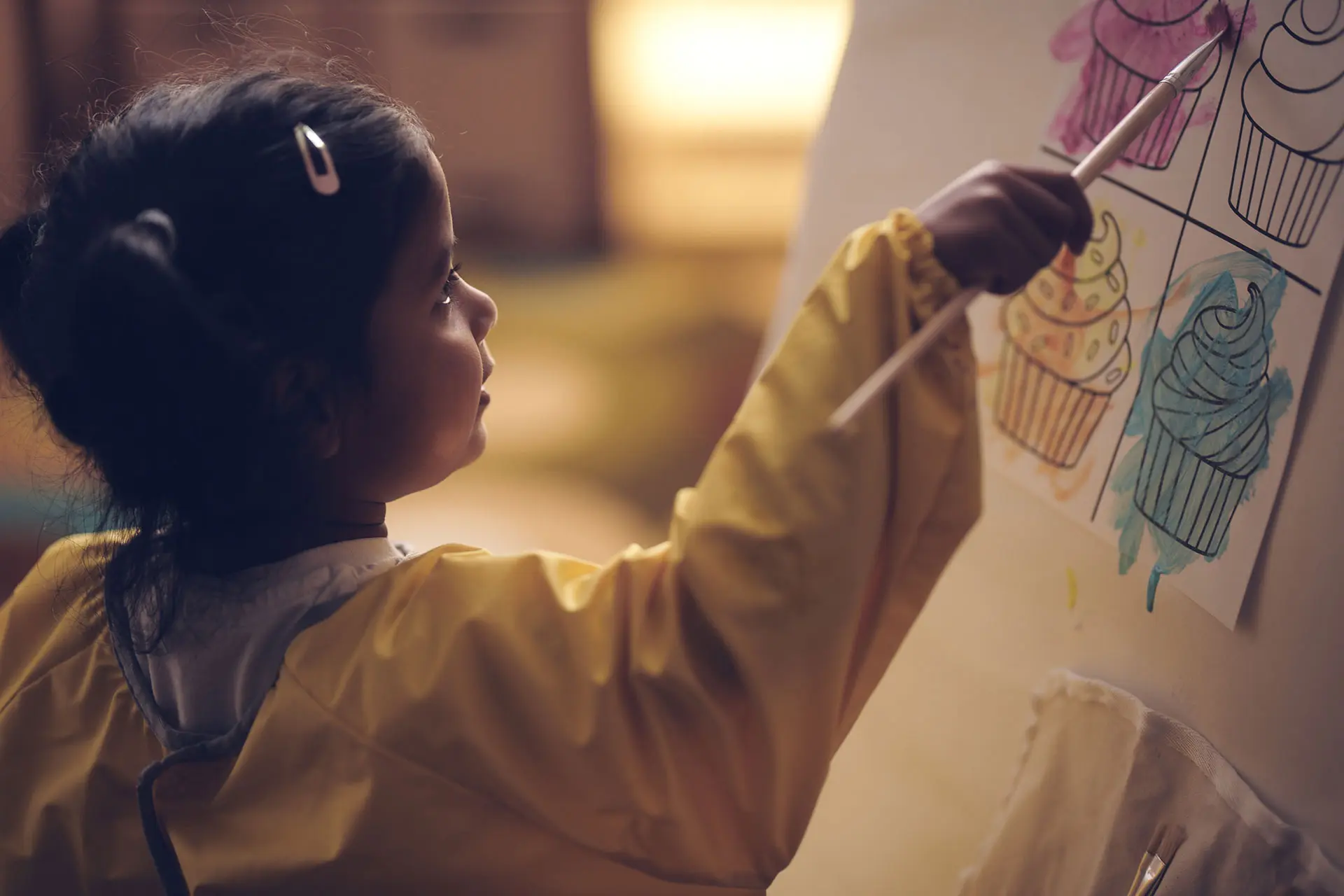 A young girl wearing a yellow smock paints colorful cupcakes on paper with a brush, focusing intently at an easel—a scene often seen in quality early learning centres that foster creativity and self-expression.