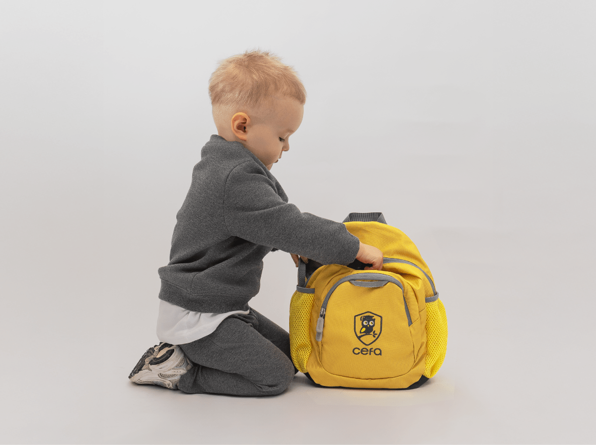 A young child with short blonde hair, dressed in a grey outfit, kneels on the floor and reaches into a bright yellow backpack with a "cefa" logo—perfect for curious minds exploring early learning centres—set against a plain white background.
