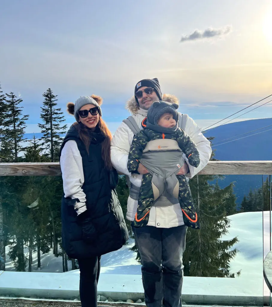 A family of three poses outdoors on a snowy mountain. The woman wears a black and white jacket and sunglasses, the man wears a white coat and beanie, and the baby is in a front carrier—perfect for parents seeking early learning centres nearby. Trees and blue sky are in the background.