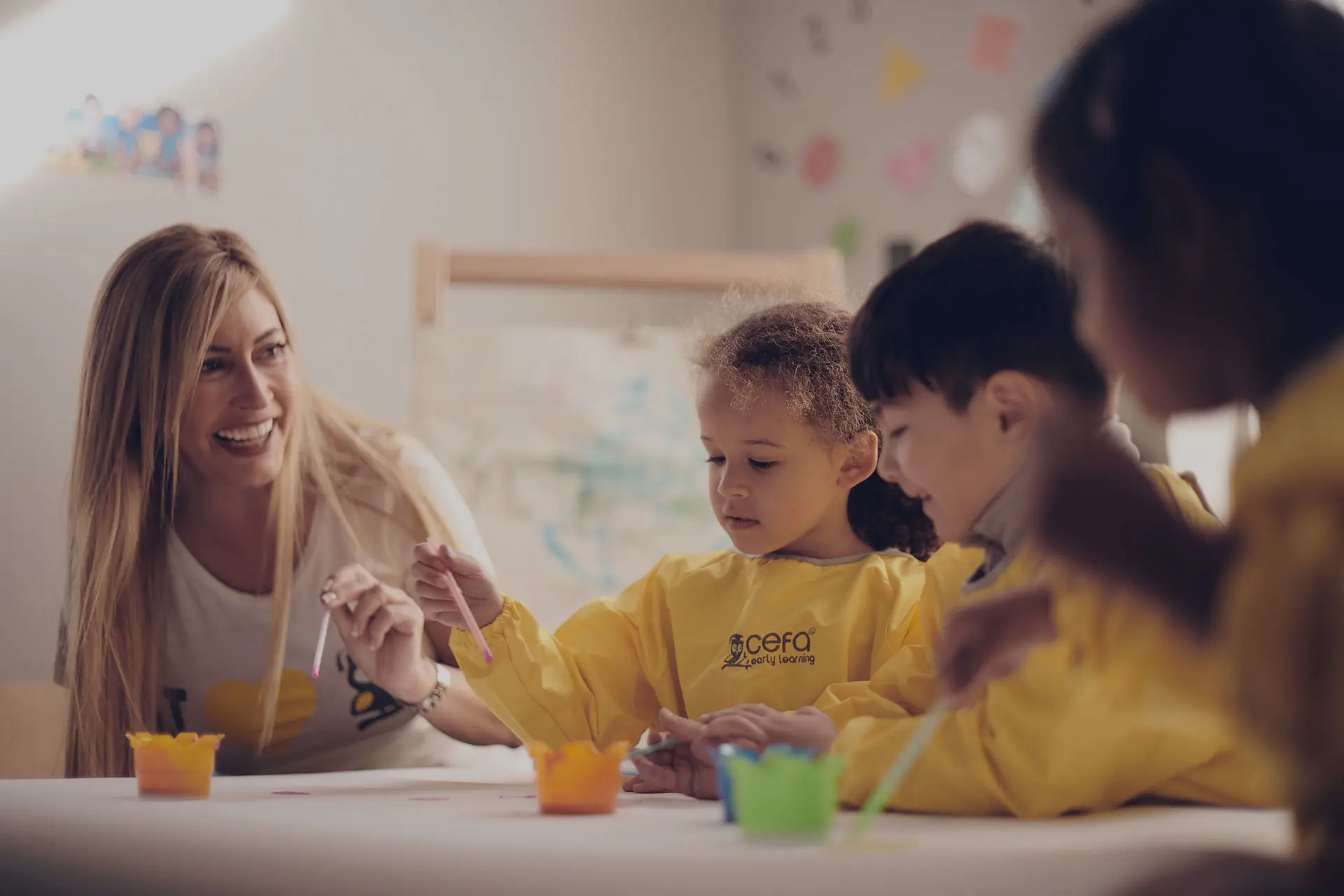 A teacher smiles as three young children in yellow smocks paint at a table in a bright classroom decorated with colorful artwork at one of the leading early learning centres.
