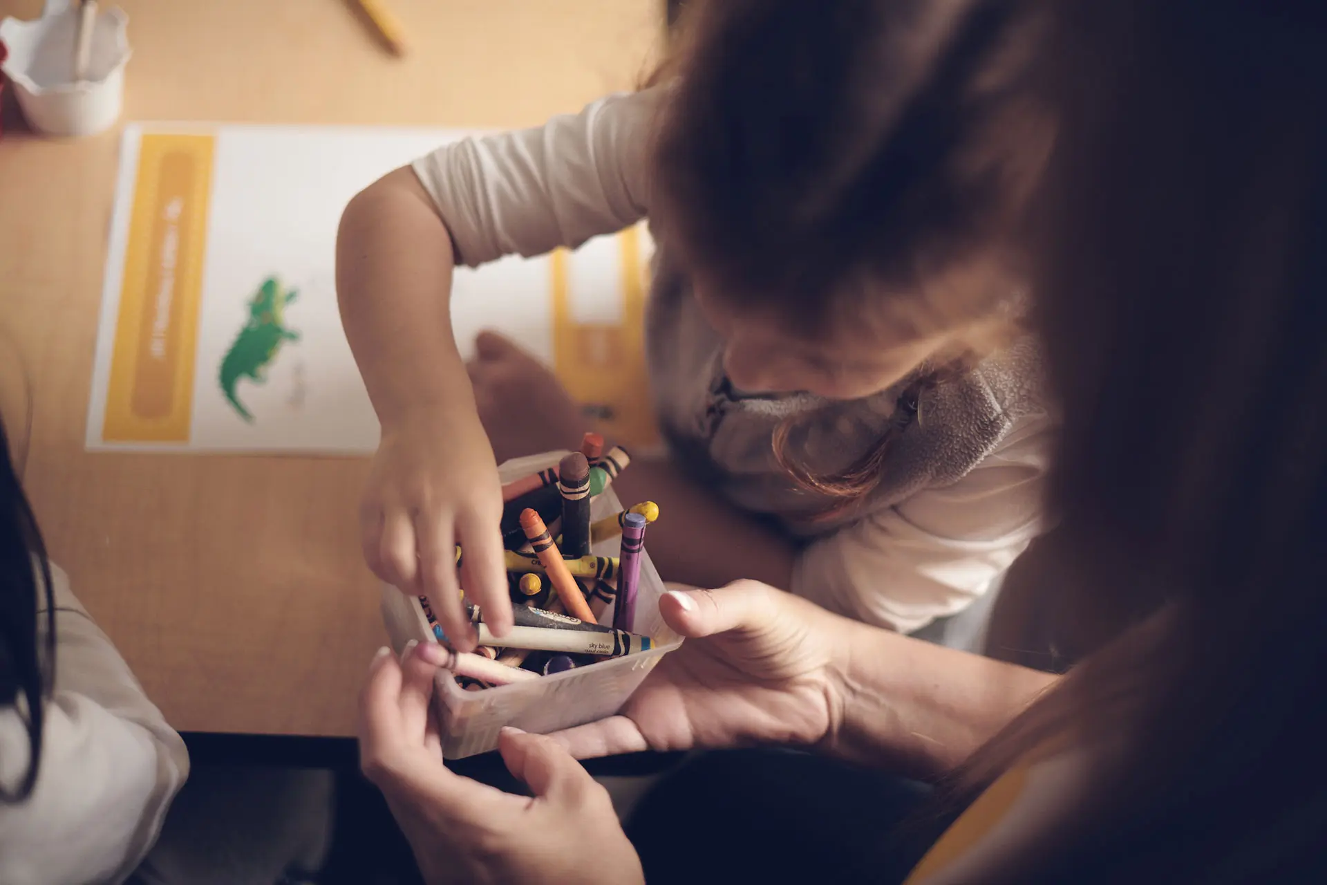 A young child reaches into a box of crayons held by an adult at one of the early learning centres, preparing to color a worksheet featuring a green alligator on a table.