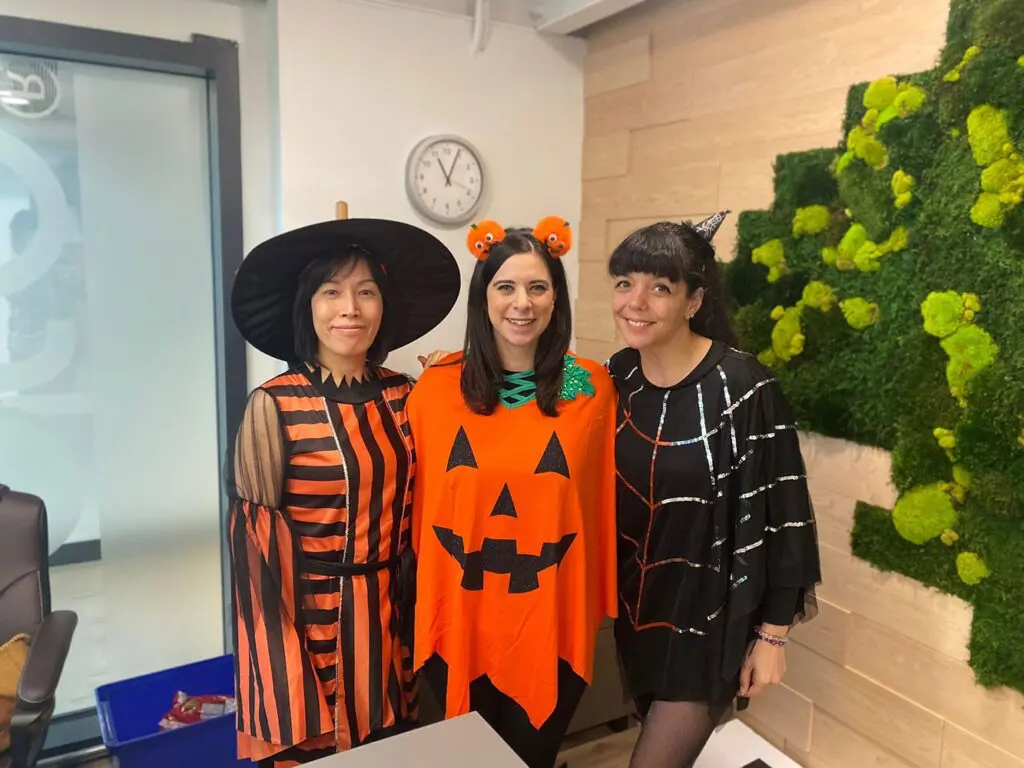 Three women in Halloween costumes—a witch, a jack-o’-lantern, and a spiderweb—stand smiling in an office with a clock and green wall decor, capturing the festive spirit often found at early learning centres and preschool schools near me.