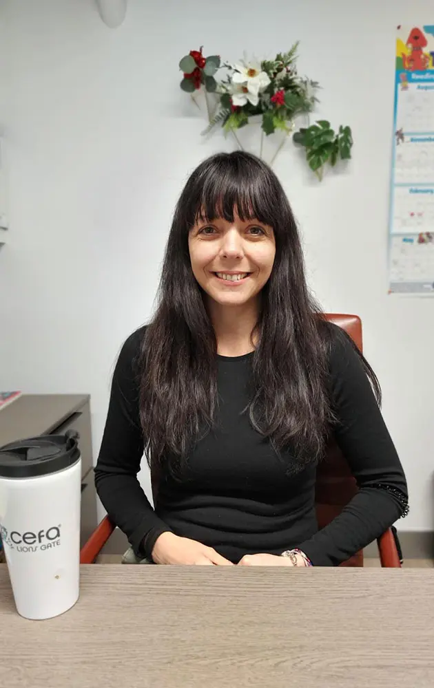 A woman with long dark hair and bangs, wearing a black top, sits at a desk smiling. There is a white travel mug on the table, a calendar on the wall, and decorative flowers behind her—perfect for early learning centres inspiration.