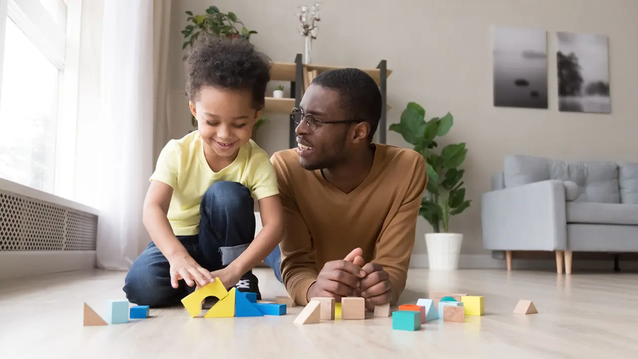 A man and a child are lying on the floor in a bright living room, smiling and playing with colorful wooden blocks together, just like activities at early learning centres.