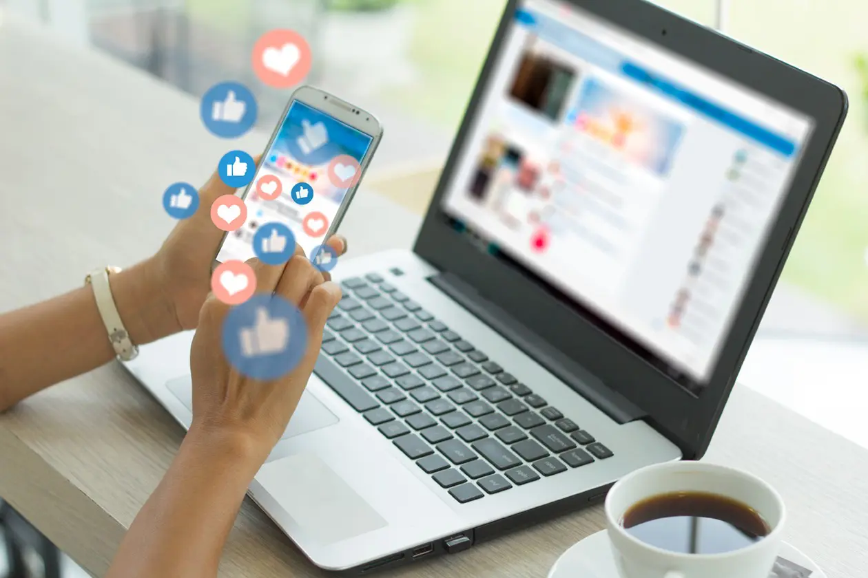 A person holds a smartphone with floating social media icons above it, while working on a laptop at a desk. A cup of coffee is nearby. The laptop screen shows courses on early childhood education and related resources.