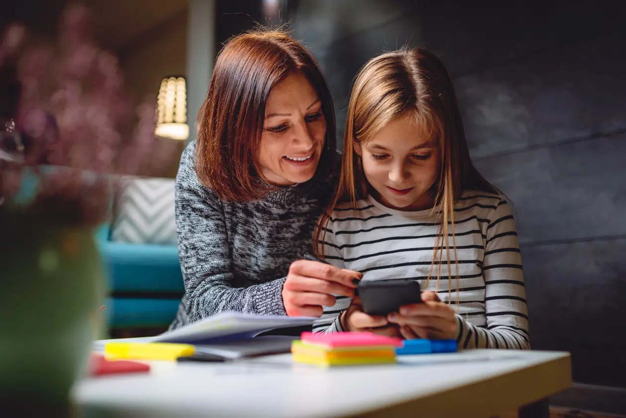 A woman and a young girl sit at a table with notebooks and colorful sticky notes. The woman smiles and points at the smartphone the girl is holding, both appearing engaged and happy—like learning together at early learning centres.