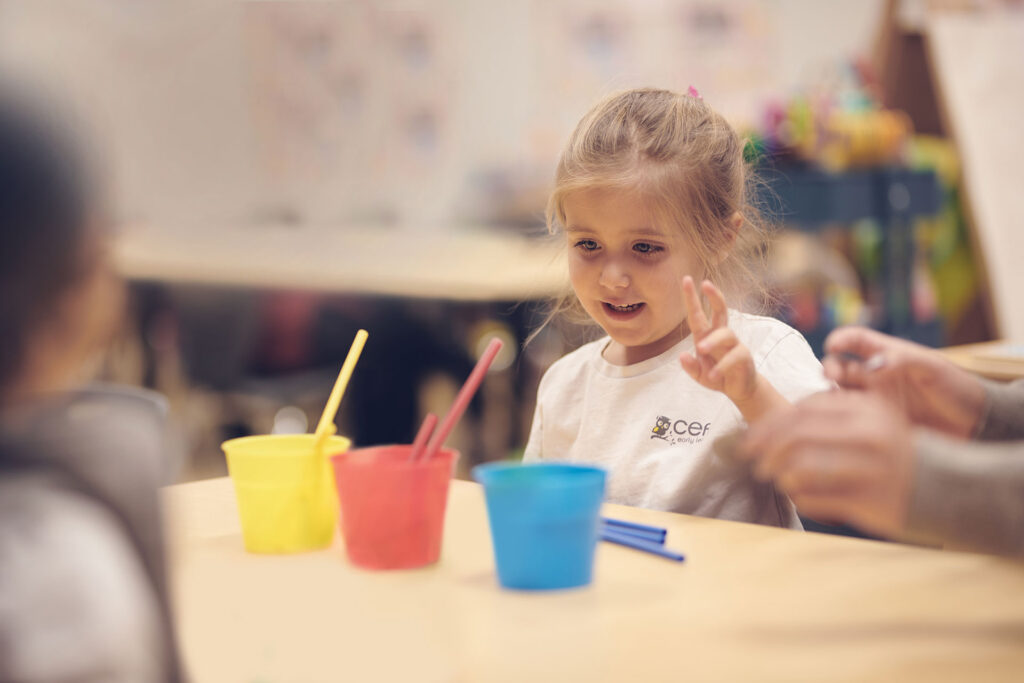 A young girl sits at a table in an early learning centre classroom, smiling and looking at colored cups with straws. An adult's hands are nearby, helping with an activity. The background is softly blurred.