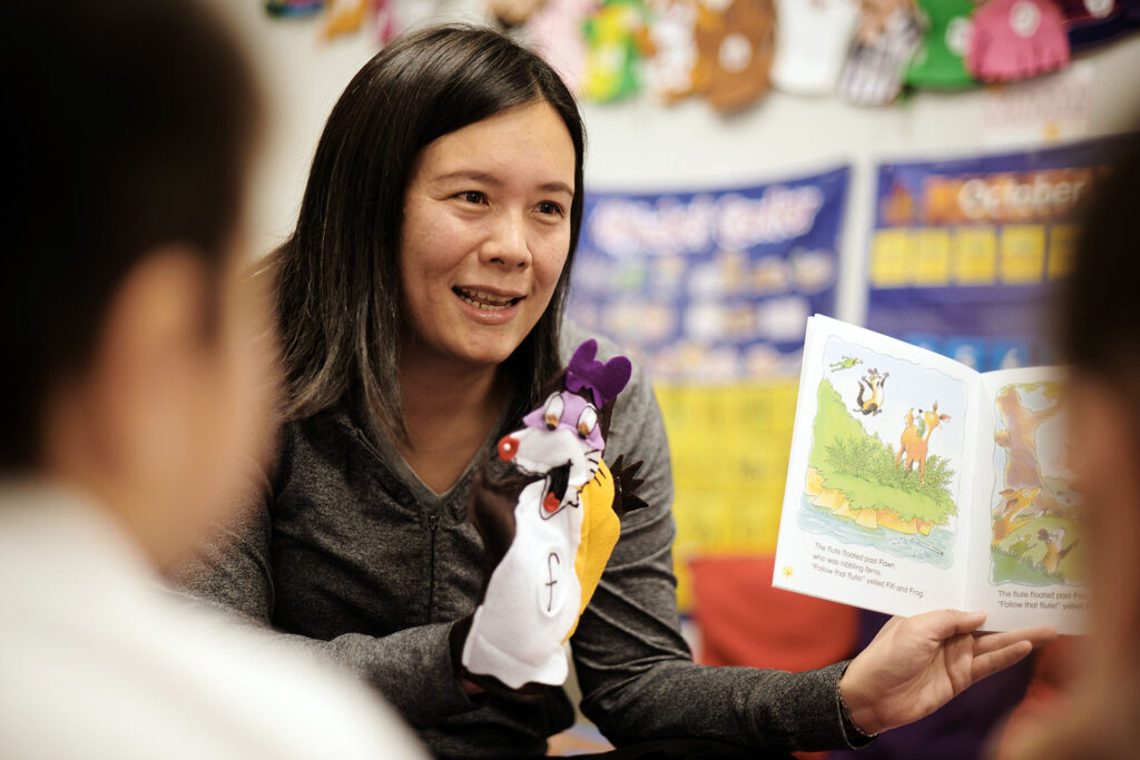 A woman holds a book open and a wolf hand puppet while reading to young children in a classroom decorated with colorful posters and artwork, creating an engaging environment often found in early learning centres.