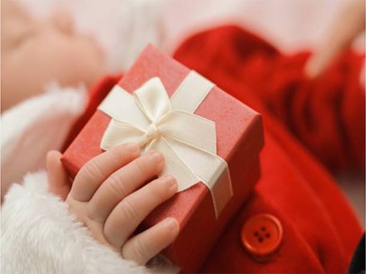 A baby dressed in red holds a small red gift box tied with a white ribbon, capturing the wonder and curiosity nurtured in early learning centres. The focus is on the baby's hand and the present.