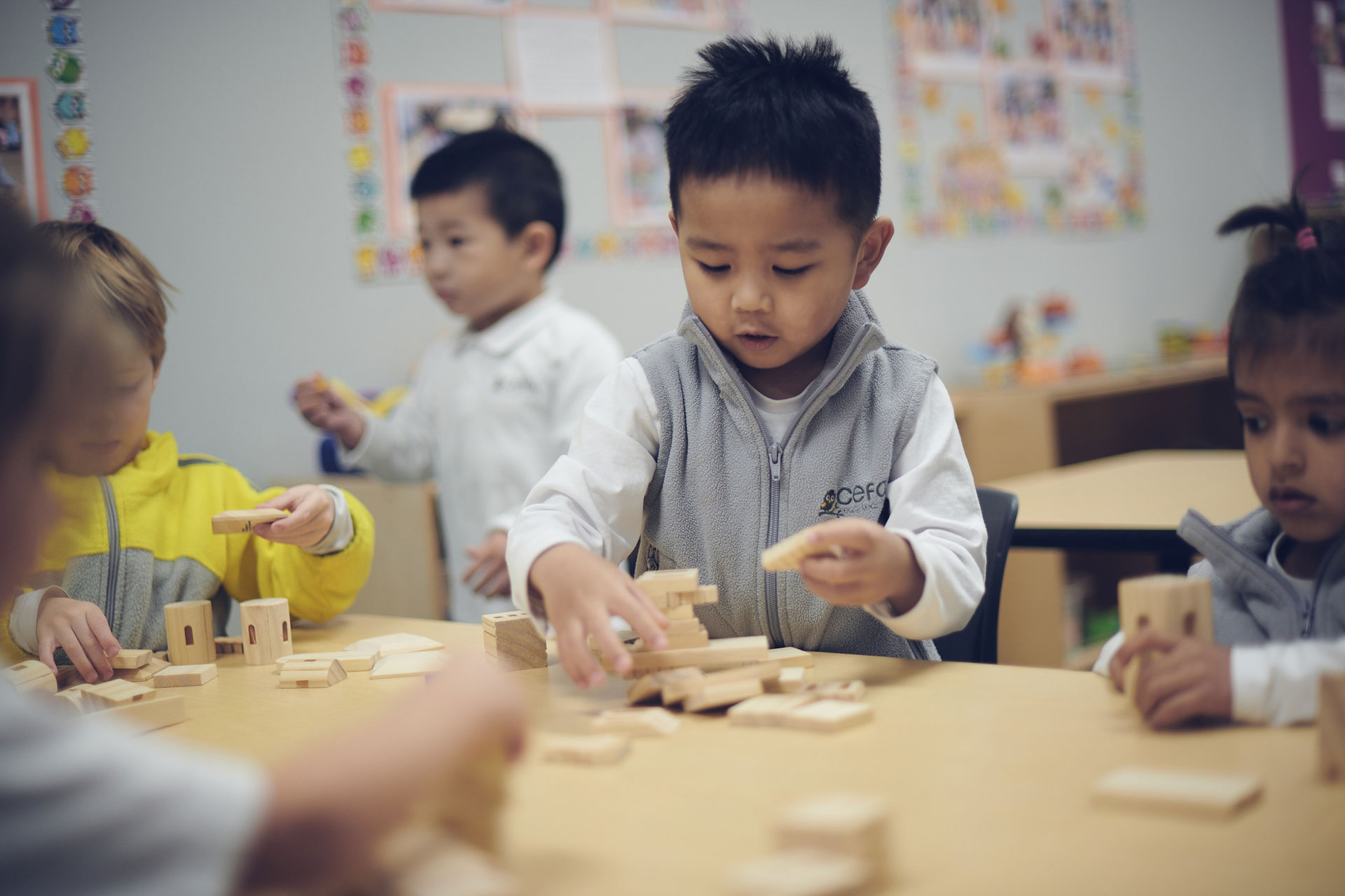 Young children sit at a classroom table, concentrating as they build structures with wooden blocks. Colorful posters decorate the wall in this lively early learning centre, fostering creativity and exploration.