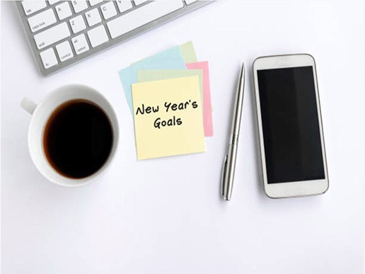 A cup of coffee, a silver pen, a smartphone, sticky notes with "New Year’s Goals"—including finding preschool schools near me—and part of a computer keyboard on a white desk.