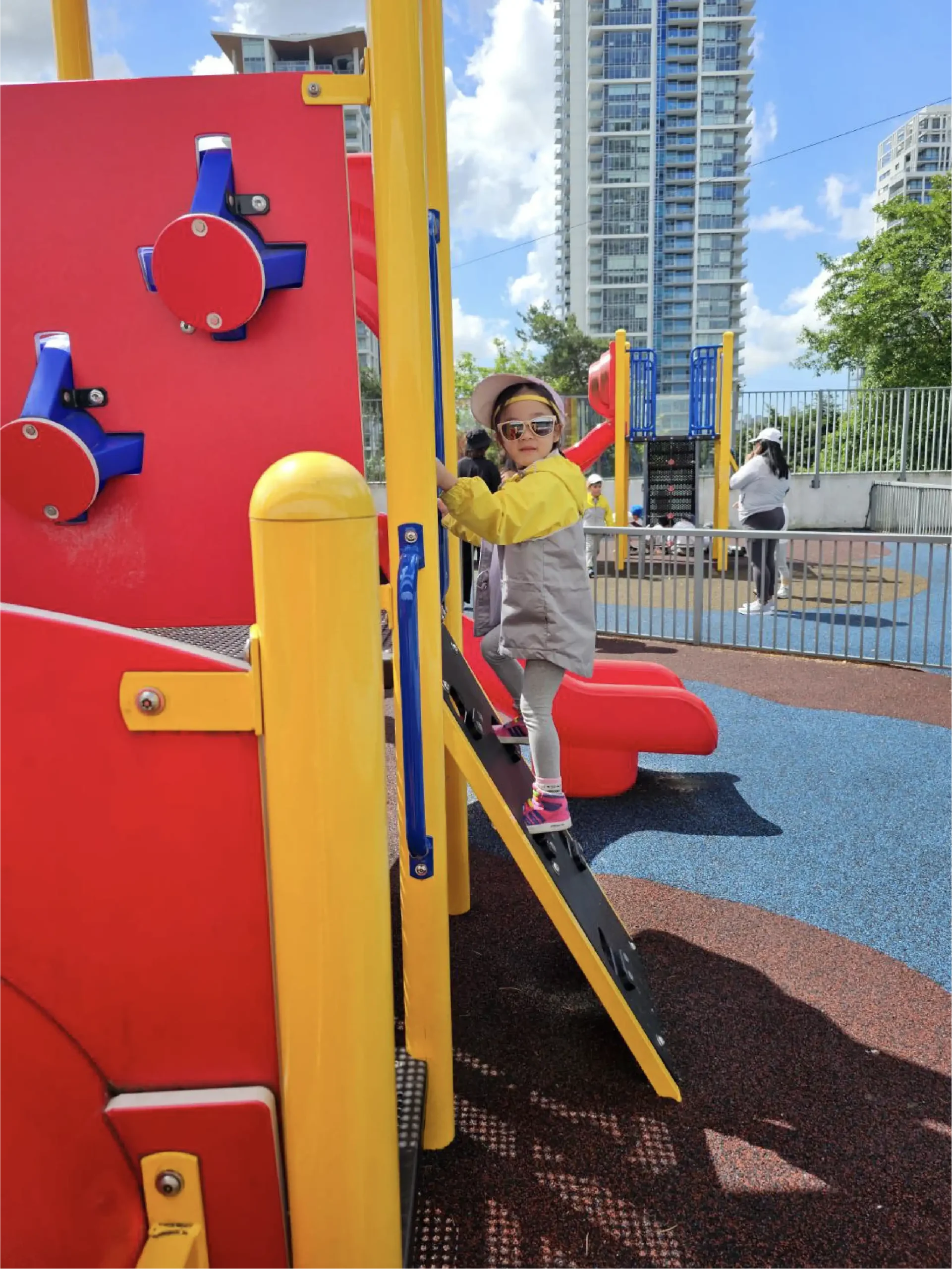A young child wearing sunglasses and a yellow jacket climbs up a play structure at a colorful playground near early learning centres, with tall buildings and a blue sky with clouds in the background.