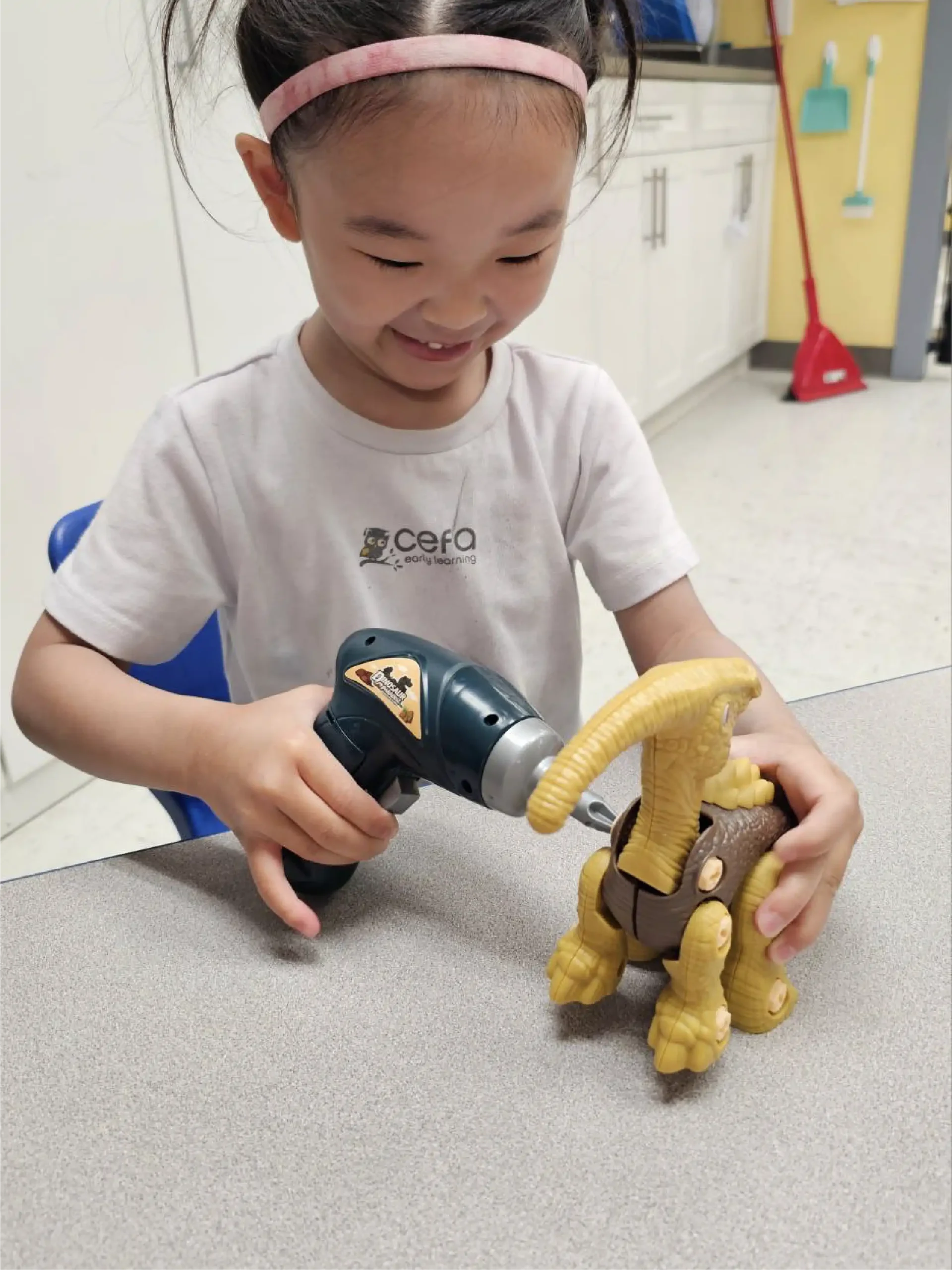 A young girl smiles while using a toy drill on a yellow dinosaur figure at a table in one of the early learning centres, creating a fun and engaging classroom setting.