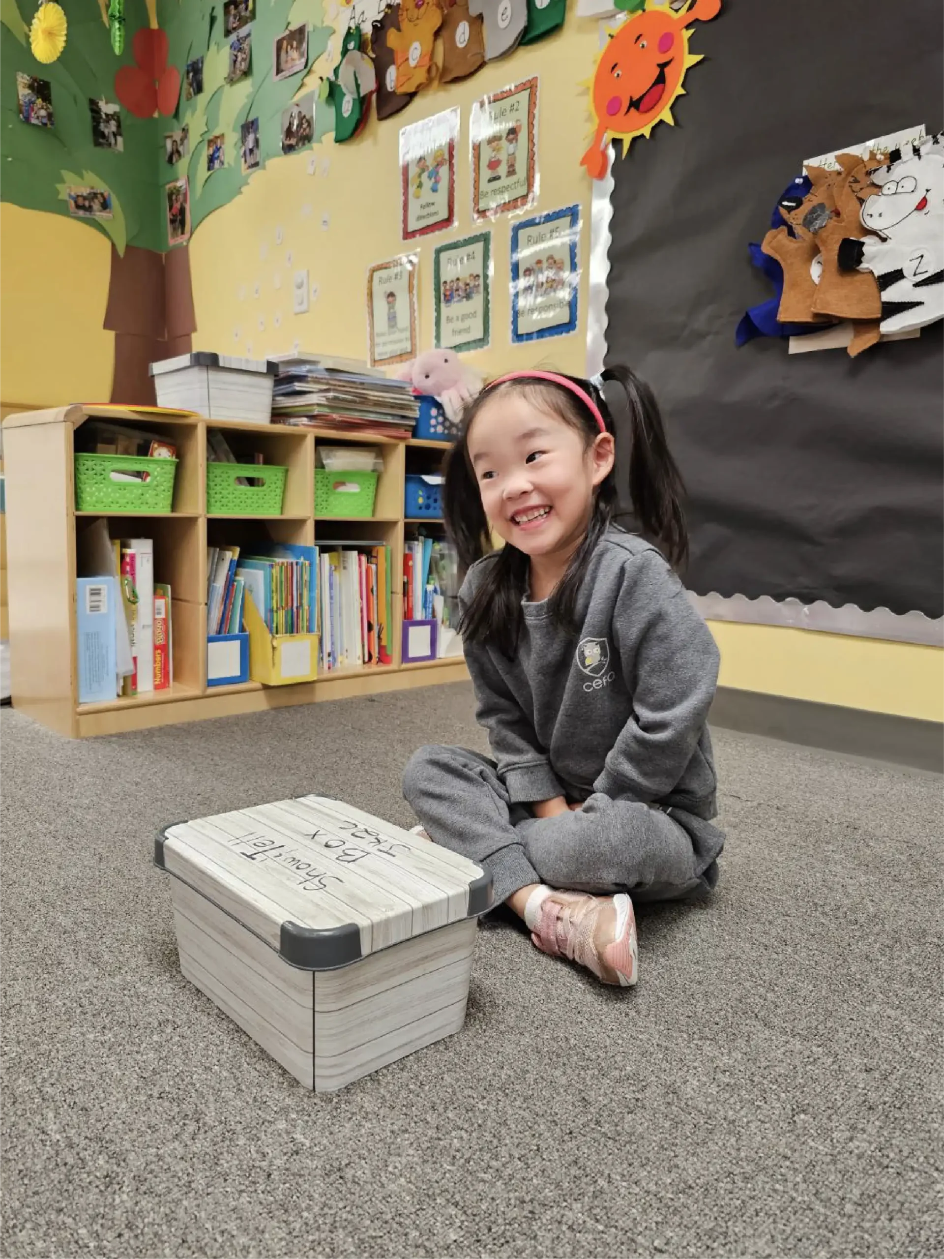 A young girl in a gray tracksuit sits cross-legged and smiling on a classroom floor beside a box of word cards. Behind her, early learning centres feature bookshelves, colorful decorations, and children's artwork on the walls.