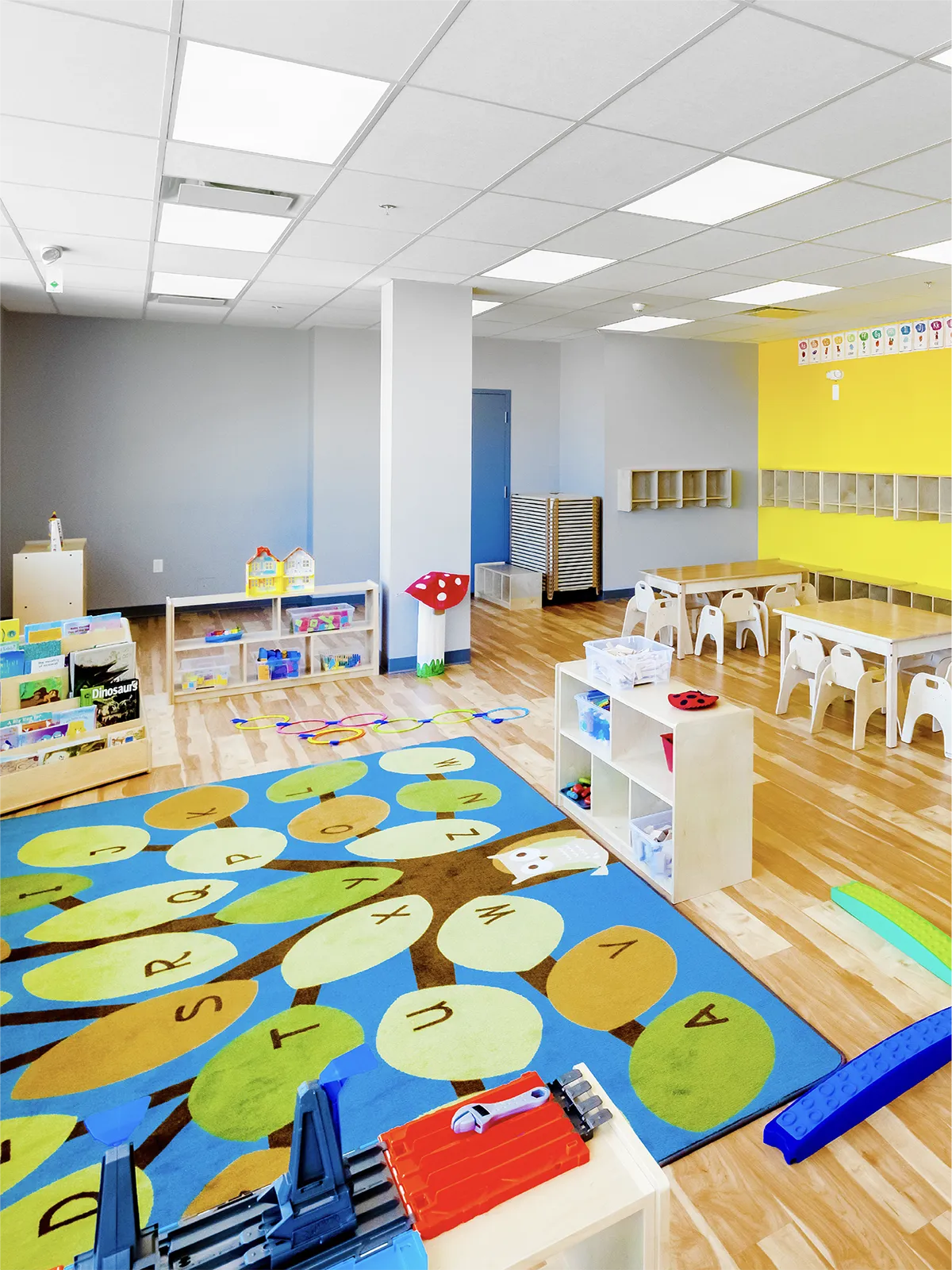 Bright, modern early learning centre preschool classroom with shelves of toys, small tables and chairs, cubbies, and a colorful alphabet rug on a wooden floor. Walls in gray and yellow feature letters above the cubbies for junior kindergarten-age children.