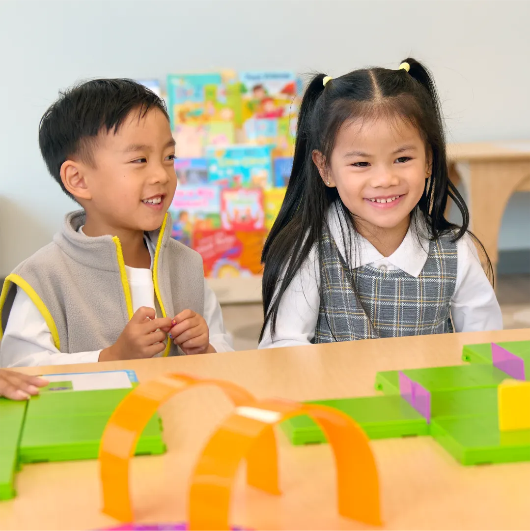 Two young children sit at a table, smiling and playing with colorful building toys in a bright classroom. Early learning centres like this, often found in preschools close to me, foster creativity and fun through hands-on activities.