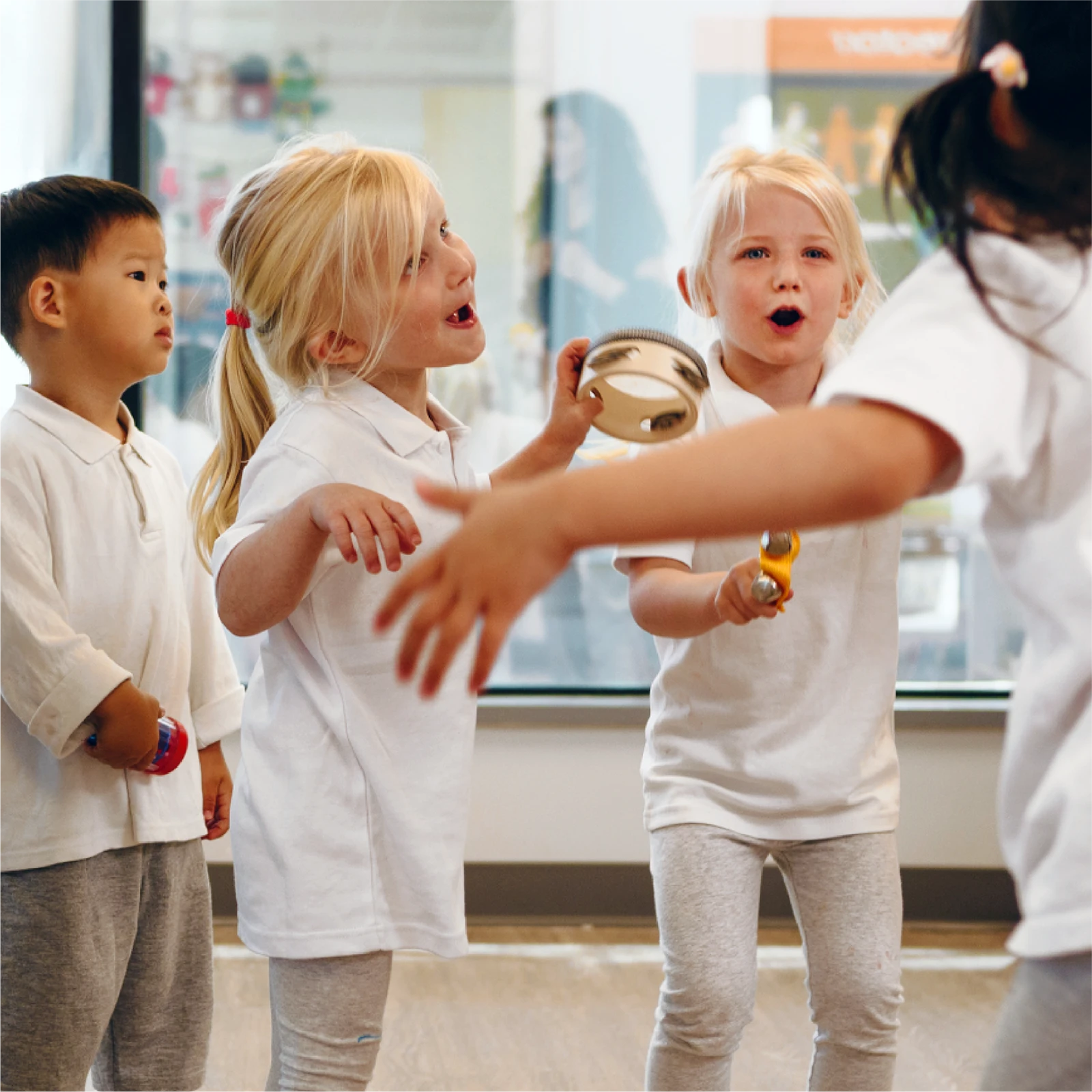 Children wearing white shirts play musical instruments and sing together indoors at one of the early learning centres. Two girls enthusiastically play tambourines, while another child holds a shaker and a fourth child’s arms are visible in the foreground.
