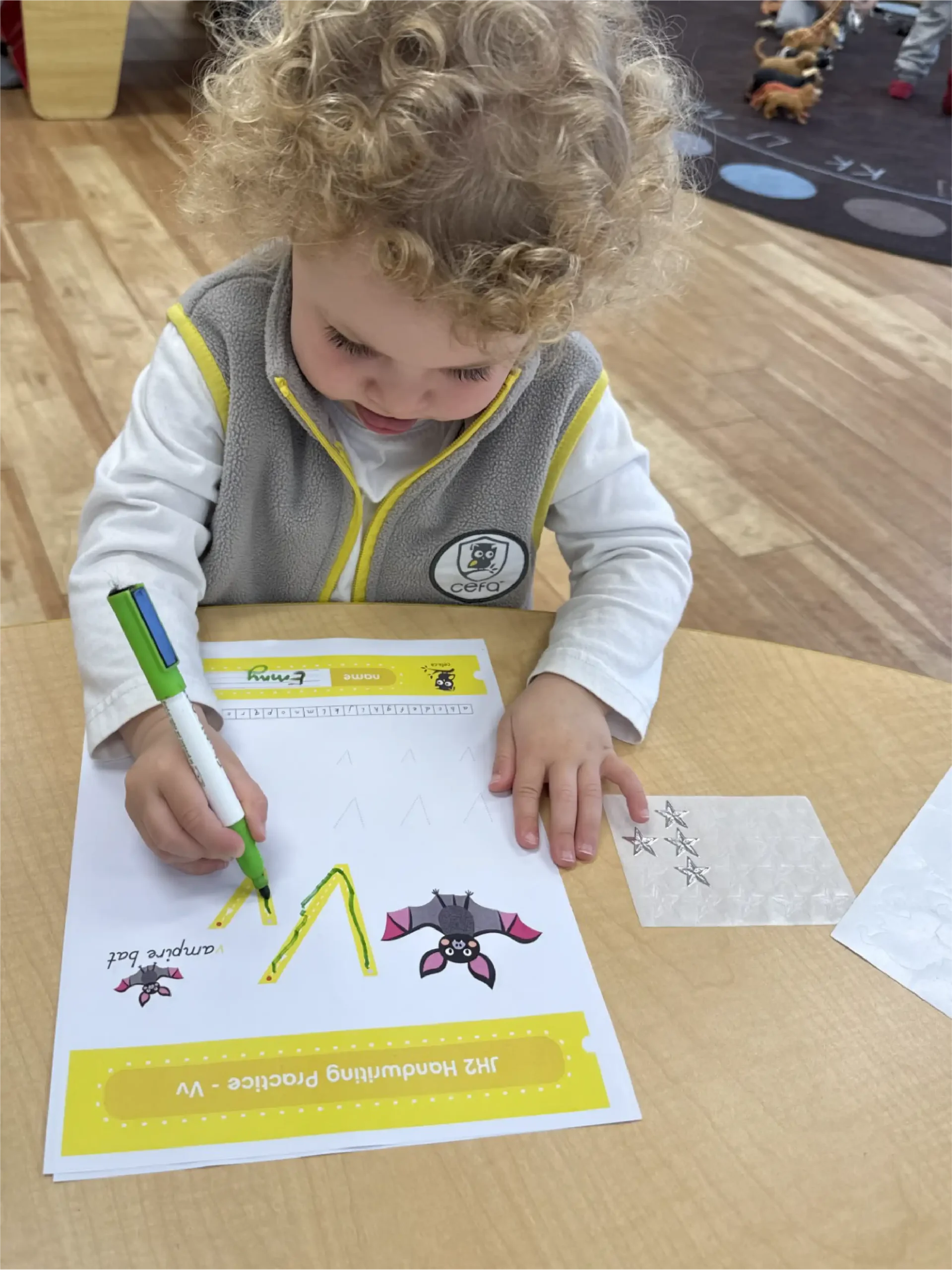 A young child with curly blonde hair uses a green marker to trace the letter "V" on a worksheet at a classroom table, enjoying handwriting practice often found in quality early learning centres.