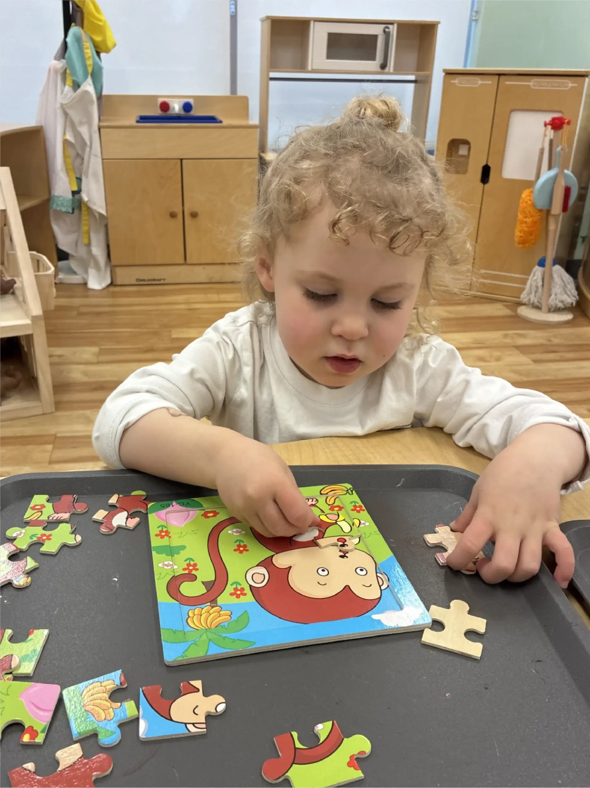 A young child with curly hair sits at a table, focused on assembling a colorful animal-themed jigsaw puzzle in an early learning centre classroom filled with wooden shelves and toys in the background.