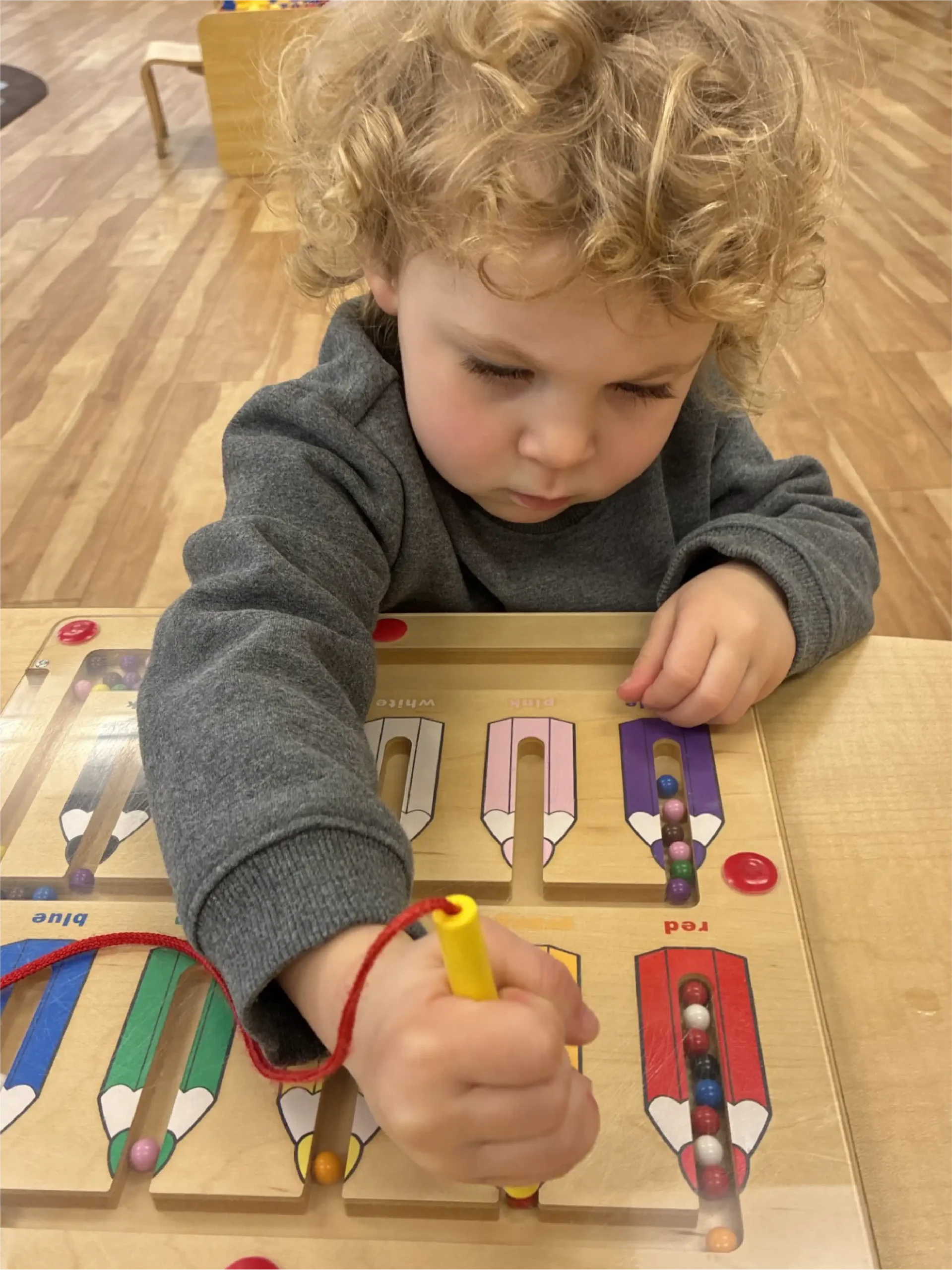 A young child with curly blonde hair concentrates while using a yellow magnetic wand to move colored beads through a wooden puzzle in an early learning centre classroom setting.
