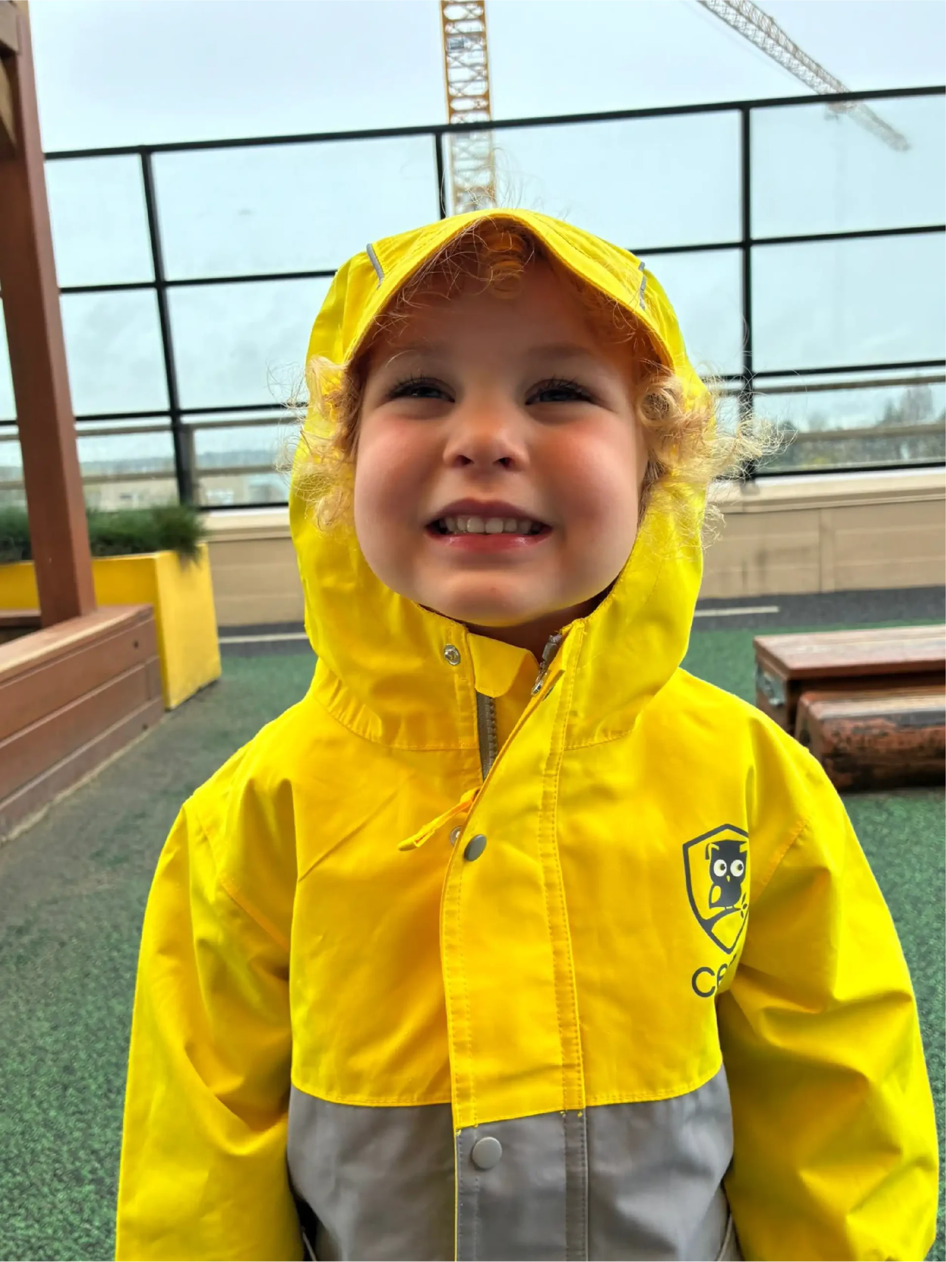 A smiling child with curly blond hair wears a bright yellow raincoat, standing outside at one of the early learning centres, with a wooden bench and potted plants in the background.