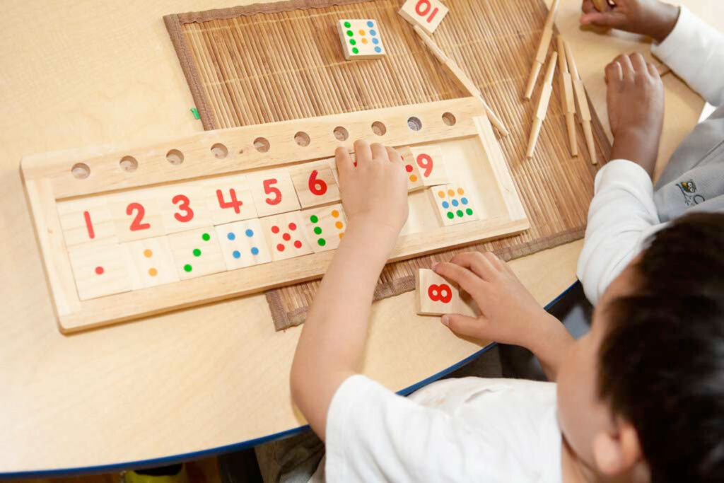 Two children sit at a table in one of the early learning centres, playing an educational counting game with wooden boards, numbered tiles, colorful dots, and clothespins to help them learn numbers and basic math skills.