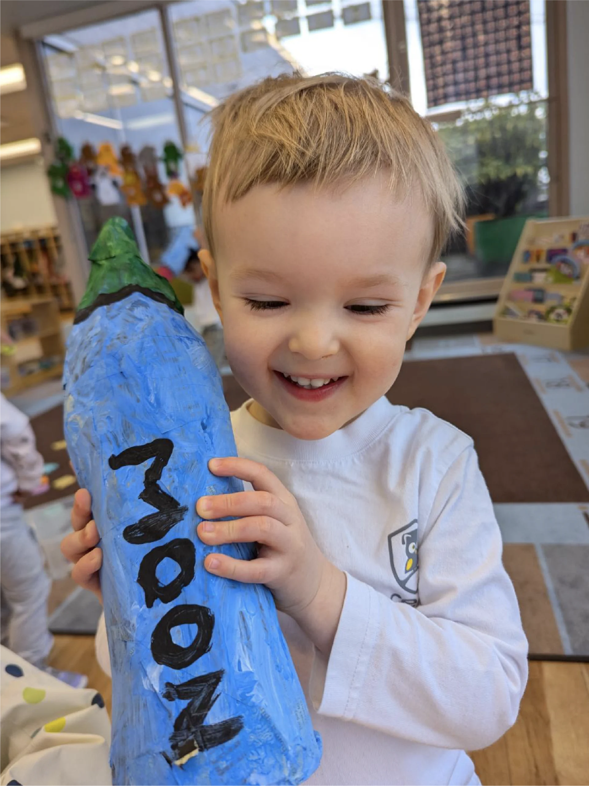A young child with short blonde hair smiles and holds a large, blue, bottle-shaped craft labeled "MOON" in black letters, standing in a brightly lit early learning centre classroom filled with artwork and toys.