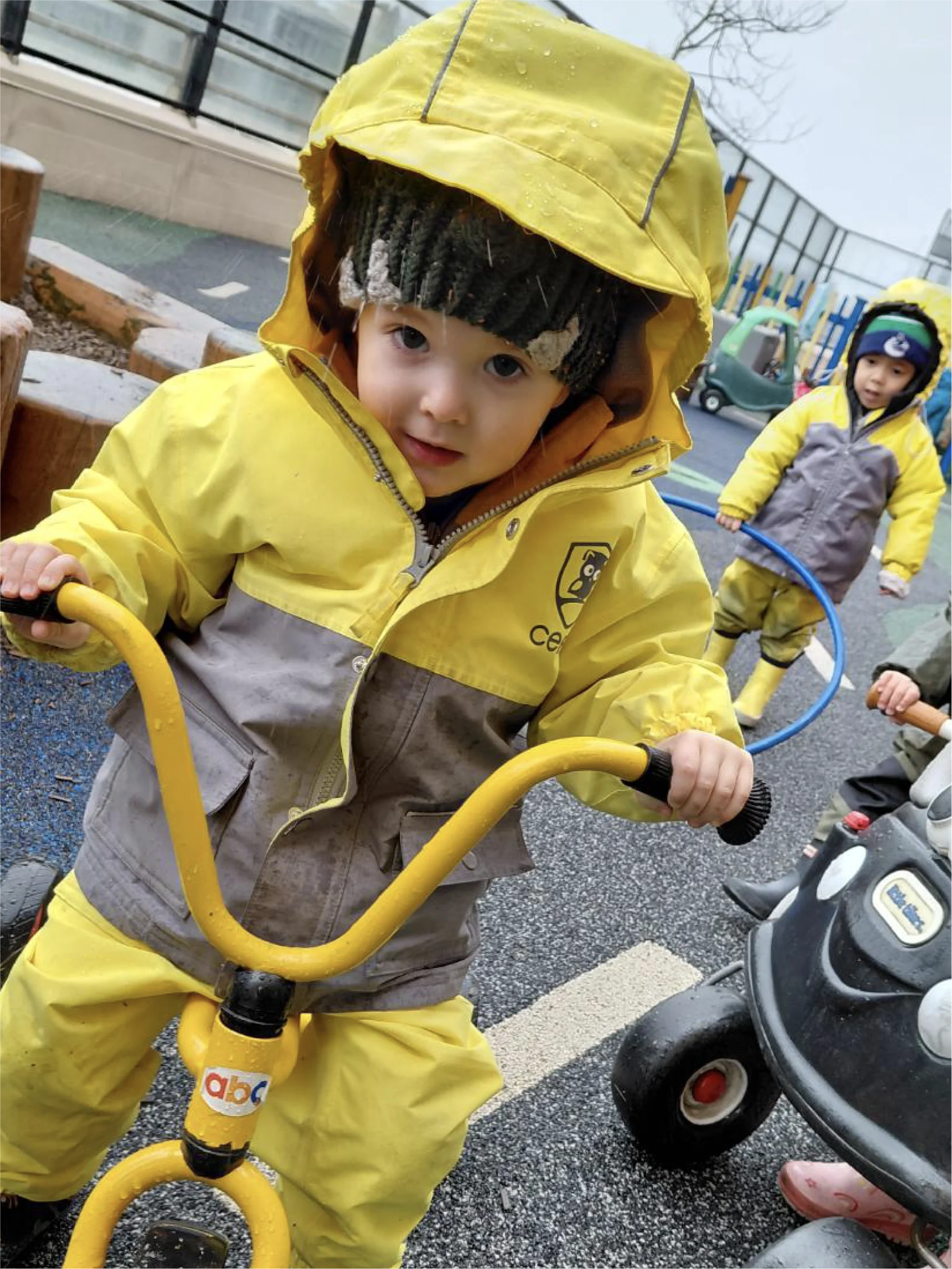 A young child in a yellow and gray raincoat rides a tricycle outdoors at one of the early learning centres, while another child in a raincoat stands by playground equipment, reflecting the engaging environments found in quality preschool schools near me.