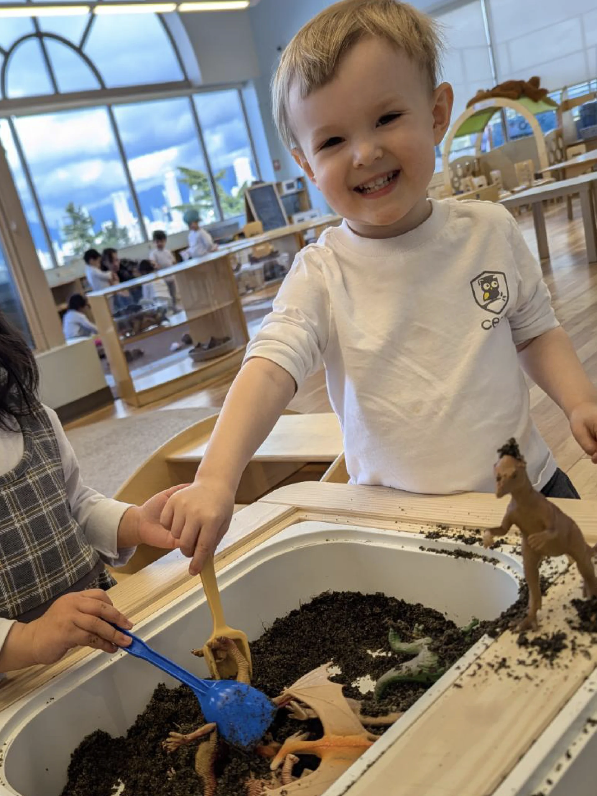 A young boy smiles while playing with toy dinosaurs and dirt at a sensory table in a bright classroom at one of the local early learning centres. Another child, partially visible, holds a blue scoop. The room has large windows and wooden furniture.