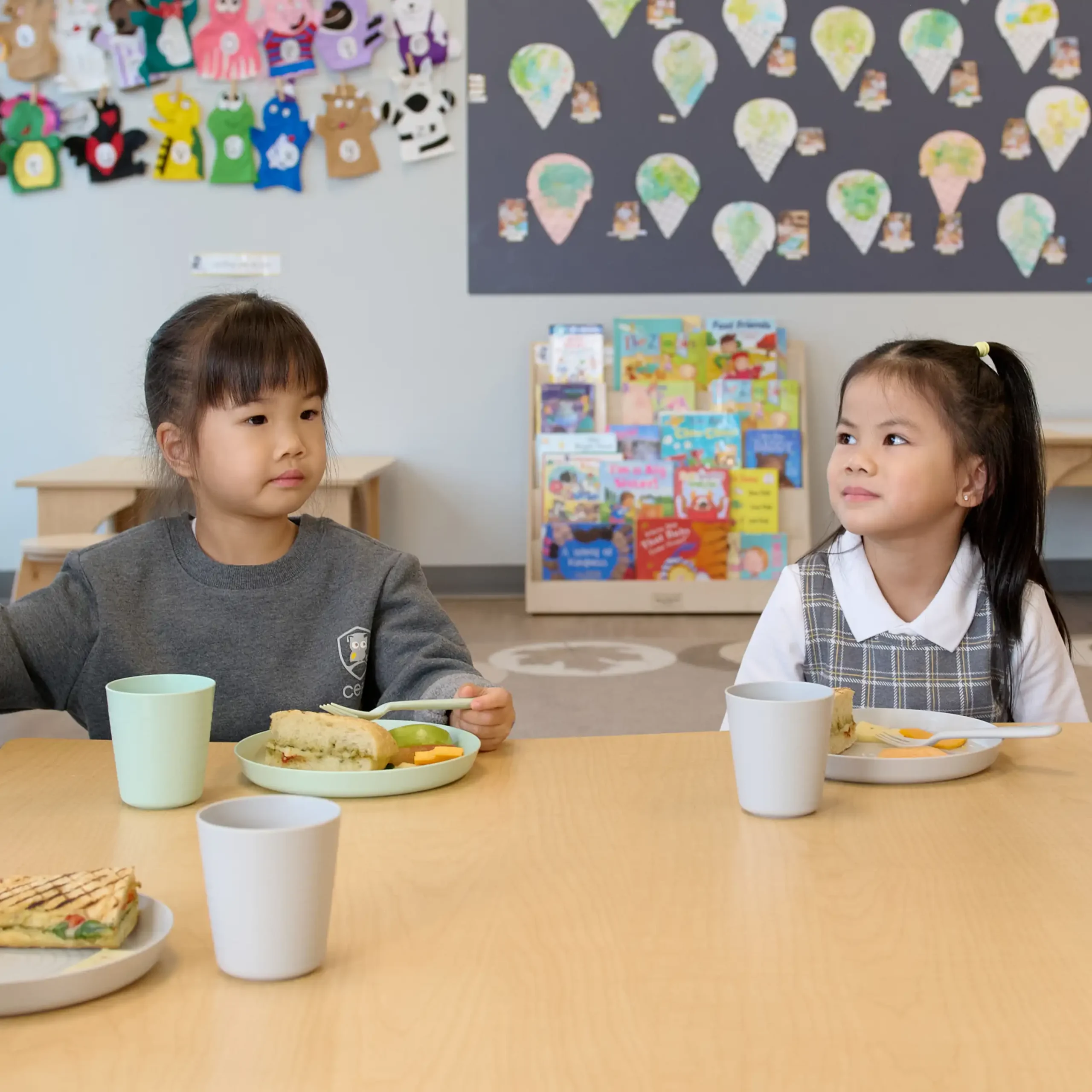 Two young girls sit at a table in a classroom, enjoying sandwiches and snacks. Surrounded by colorful books and artwork, they appear engaged—an inviting scene typical of quality early learning centres or preschools close to me.