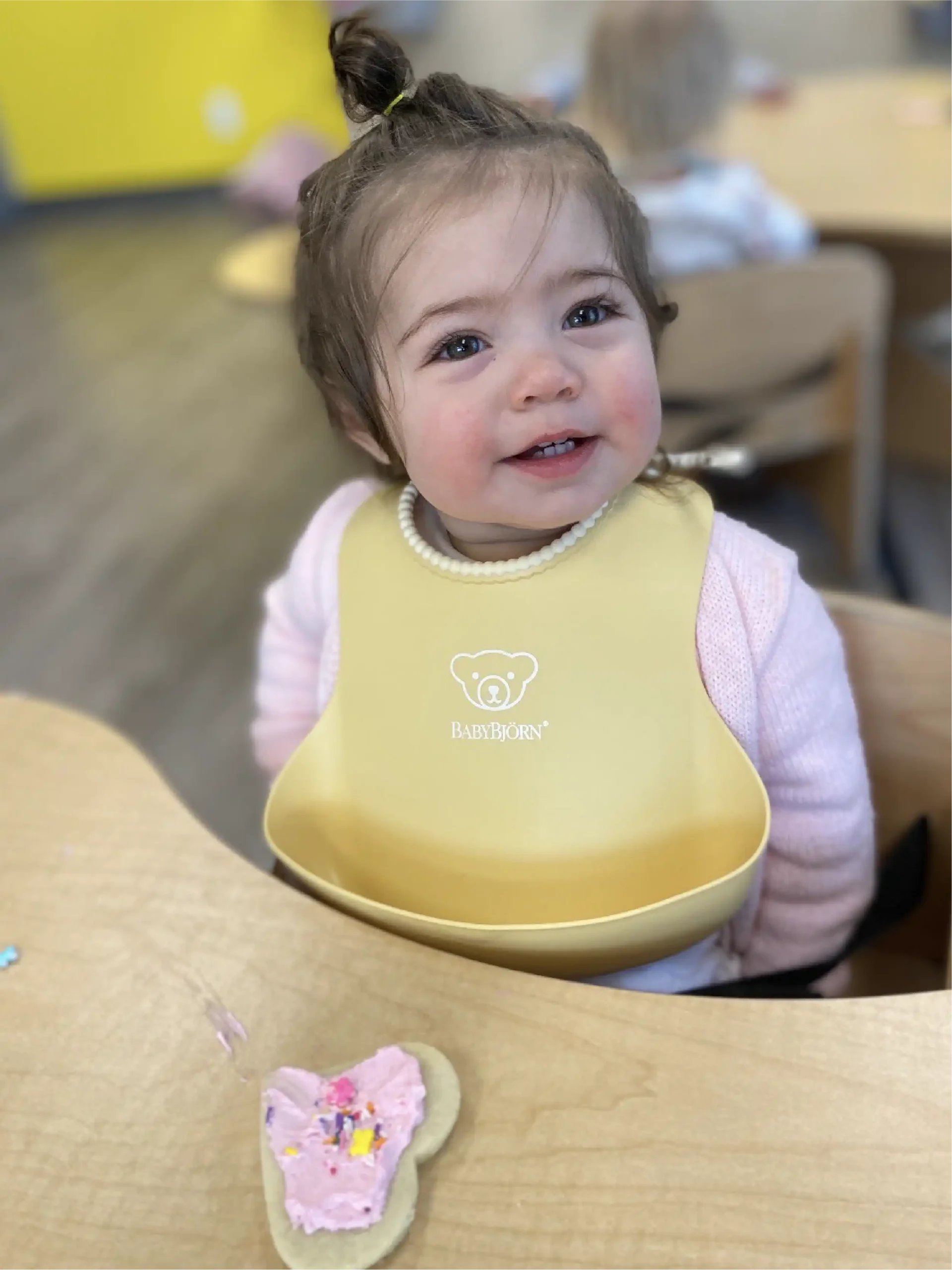 A smiling toddler wearing a yellow BabyBjörn bib sits at a table with a cookie decorated with pink frosting and sprinkles in front of her, capturing the joyful atmosphere often found in early learning centres. Other children are blurred in the background.