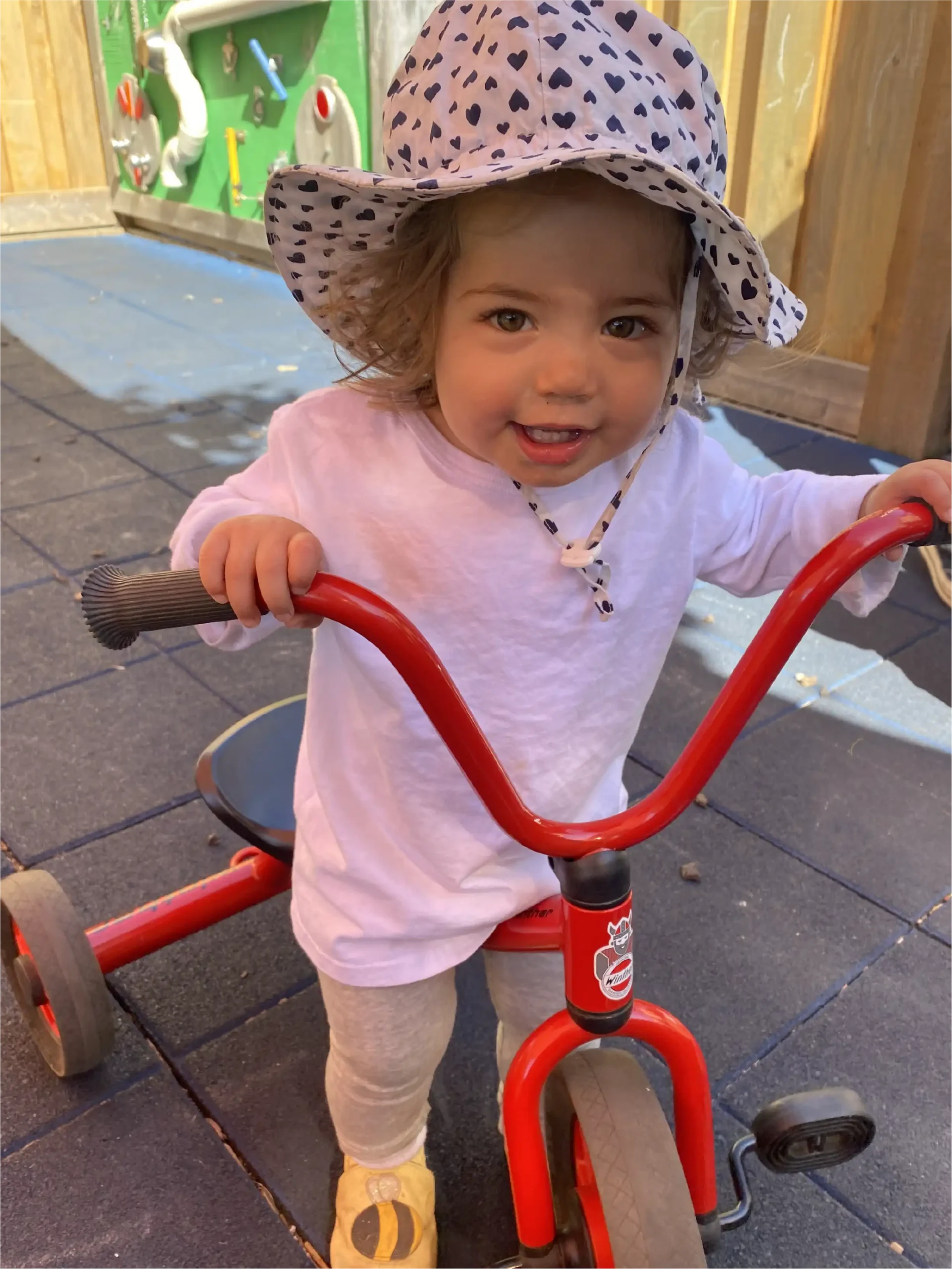 A young child wearing a white shirt, patterned hat, and sneakers smiles while riding a red tricycle on the playground of one of the early learning centres with safe rubber flooring.
