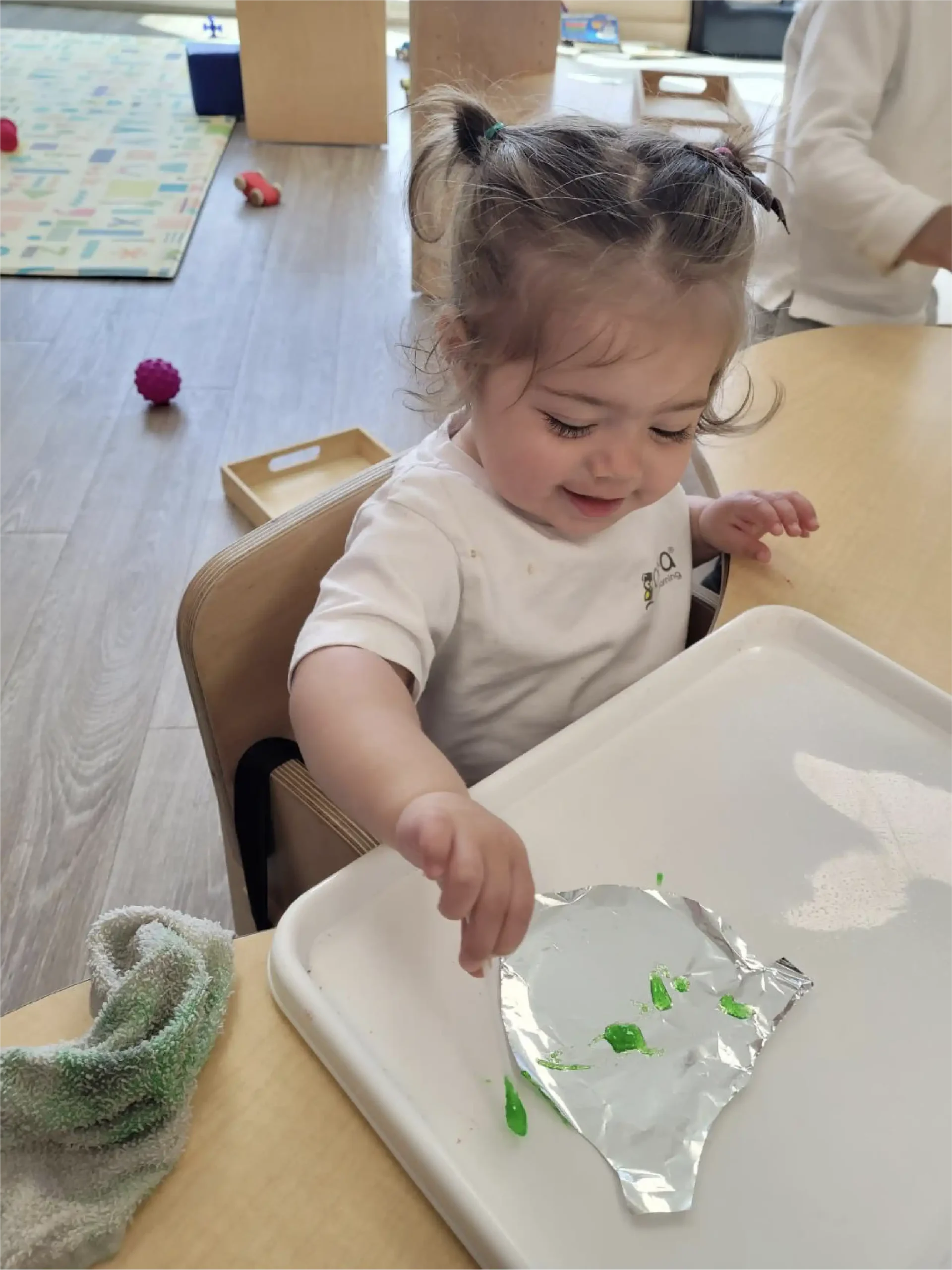 A young child sits at a table, smiling while finger painting green paint on aluminum foil placed on a white tray. A towel and toys are visible in the background, creating a lively scene often found in early learning centres.