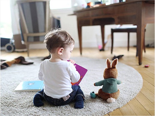 A toddler sits on a round rug reading a colorful book, with a stuffed animal bunny beside them. Sunlight fills the cozy room—much like the inviting spaces found in many early learning centres or preschool schools near me.