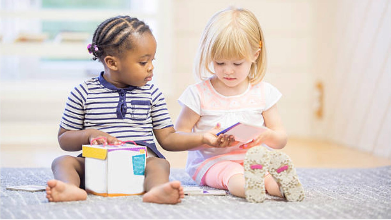 Two young girls sit on the floor together, one holding a book and the other a tablet, sharing and interacting in a bright, cozy room—an inspiring scene often found in early learning centres.