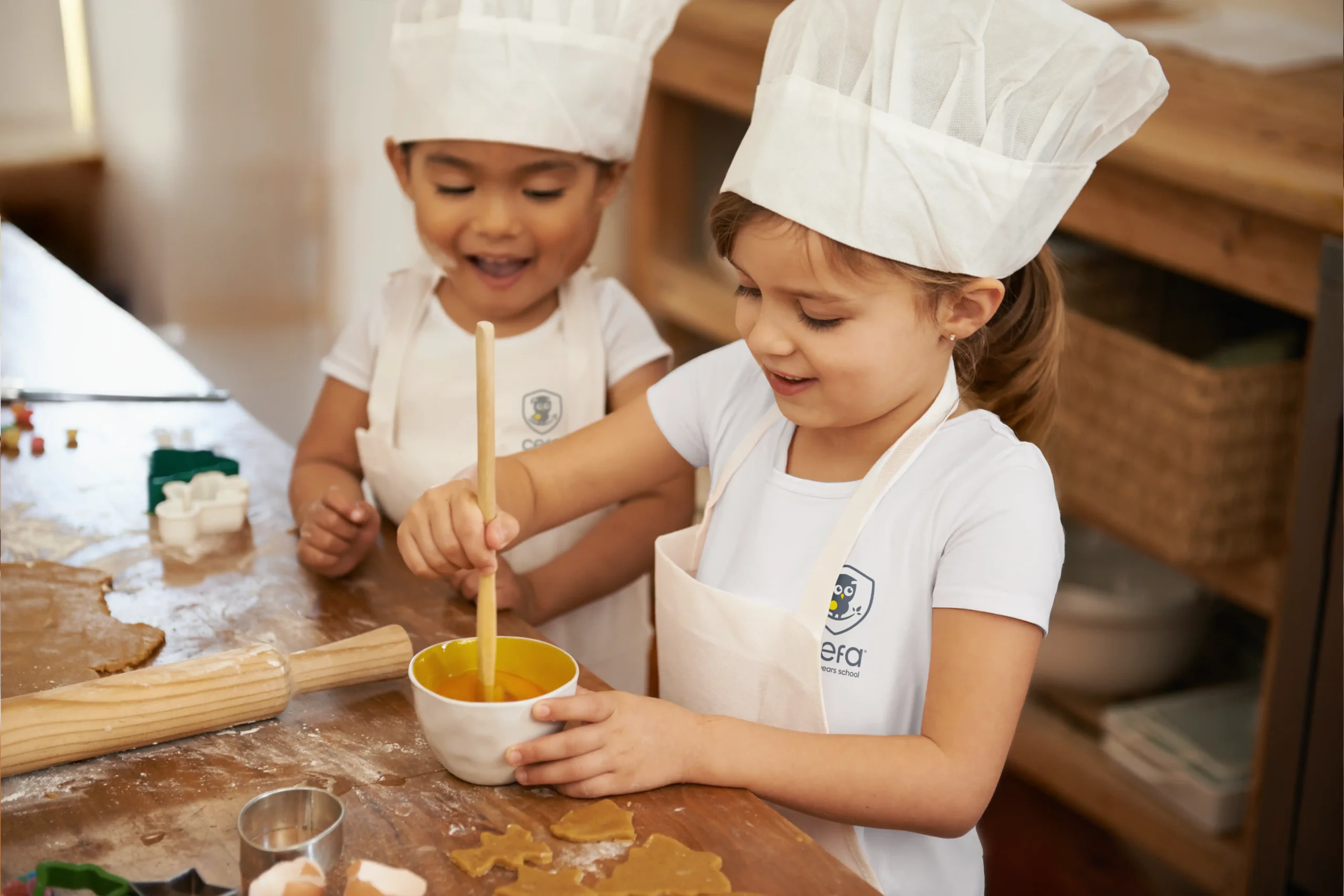 Two young children wearing chef hats and aprons smile while baking together at one of the early learning centres. One stirs a bowl with a wooden spoon; cookie cutters, dough, and baking tools are on the table in front of them.