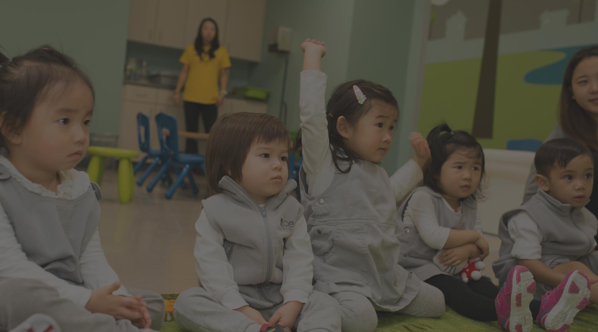 A group of young children sit on the floor in a classroom, with one child raising her hand. An adult stands nearby. The children appear attentive and engaged, reflecting the interactive environment found in top early learning centres.