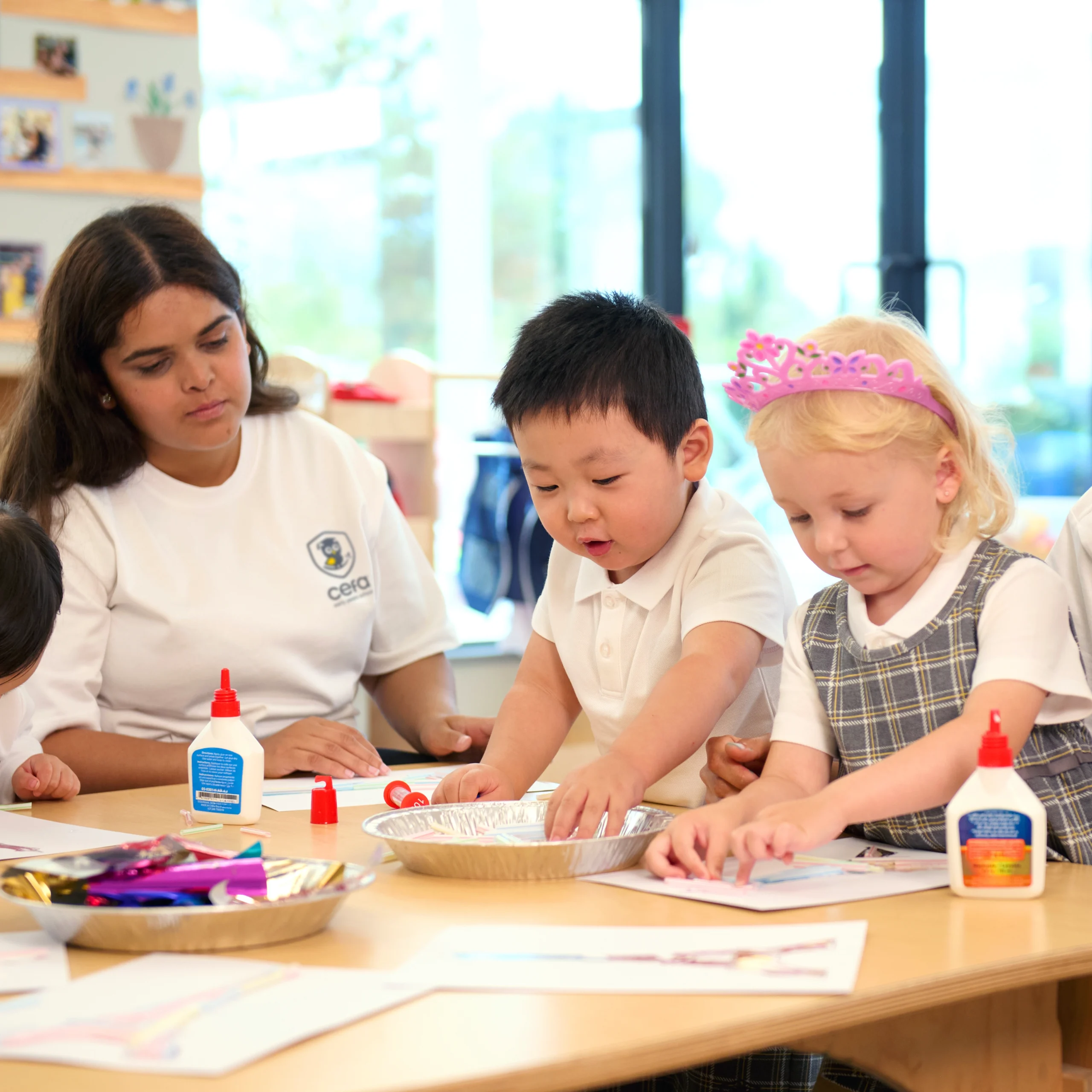 Three young children and a teacher sit at a table doing arts and crafts with glue, paper, and other materials. One girl wears a pink tiara, and the children are focused on their creative activity at one of the trusted early learning centres.