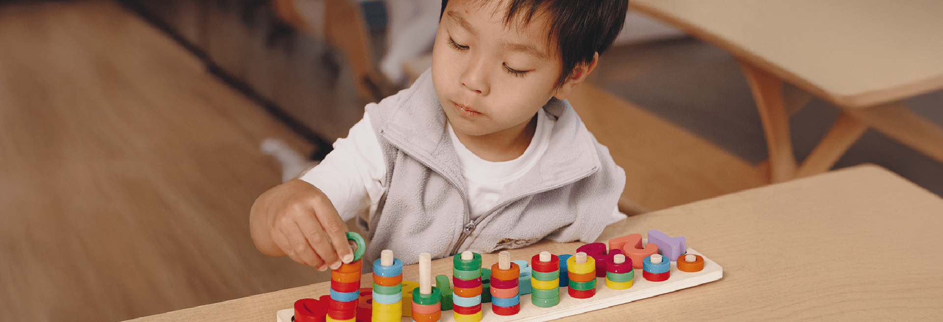 A young child sits at a table, focused on stacking colorful rings onto pegs of a wooden educational toy inside an early learning centre classroom.