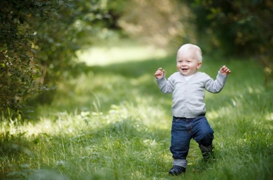 A smiling toddler with blonde hair, wearing a grey sweater and jeans, walks on a grassy path surrounded by greenery on a sunny day, embodying the joyful spirit found at early learning centres.