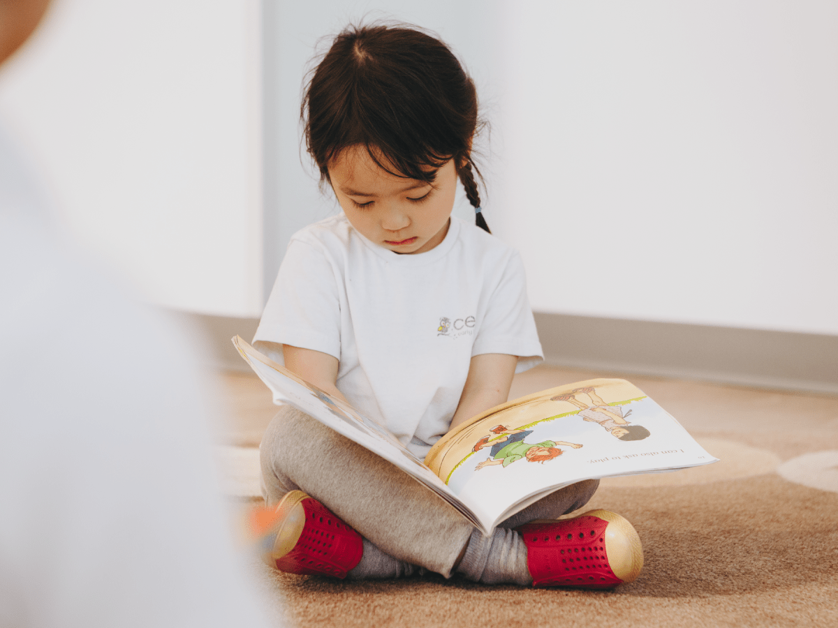 A young girl with braided hair sits cross-legged on a carpeted floor, wearing a white t-shirt and red shoes, reading an open picture book intently—an inspiring scene often found in early learning centres.