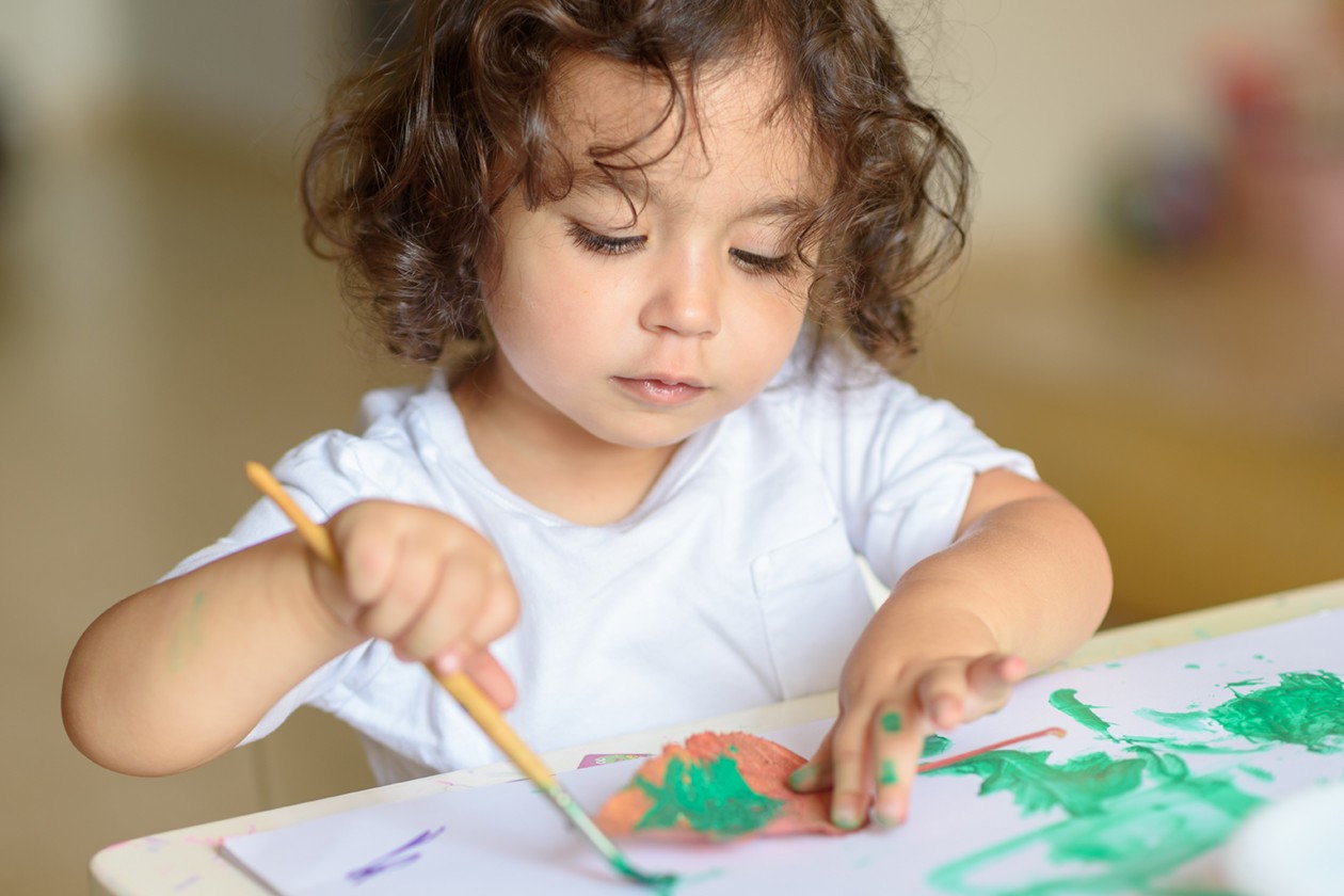 A young child with curly hair, wearing a white t-shirt, paints with a brush on paper at one of the local early learning centres. The child’s fingers and hand have green paint on them, and they appear focused on their artwork.