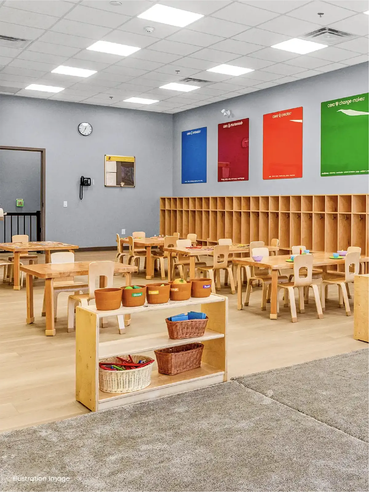 A bright, organized classroom in an early childhood care program, with small wooden tables and chairs, cubby shelves along the wall, colorful posters. Art supplies and baskets rest on a low shelf; a clock and bulletin board hang on the gray wall.
