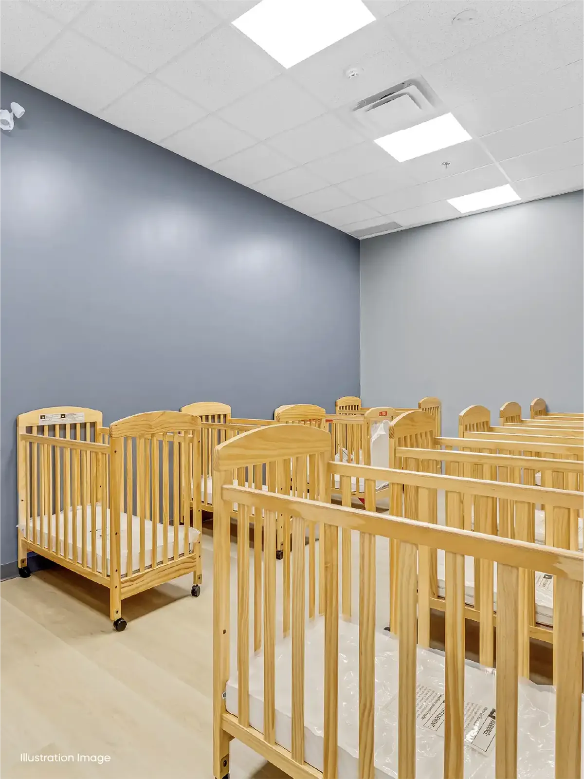 A daycare room at one of the top early learning centres, featuring several empty wooden cribs arranged in rows on a light-colored floor, gray and white walls, and bright overhead lighting.