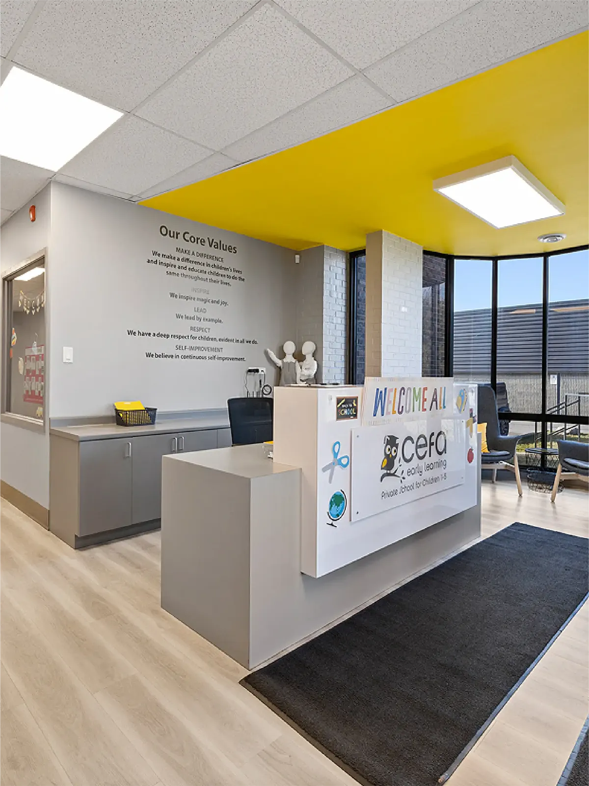 A modern daycare reception area with a gray desk, colorful welcome sign, and wall text listing core values—perfect for families exploring early learning centres. The room features a yellow ceiling, large windows, and bright lighting.