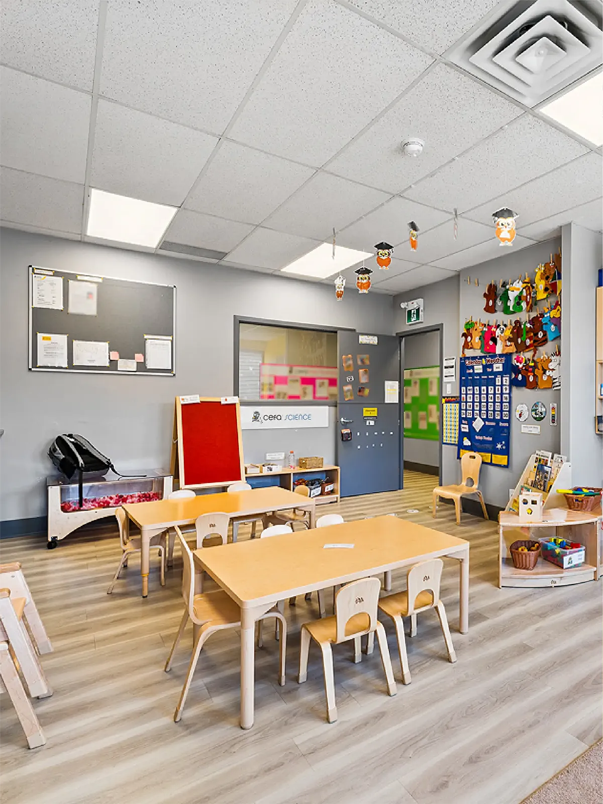 A bright, organized classroom at local early learning centres features small wooden tables and chairs, colorful wall decorations, educational materials, and children’s art displayed on the walls. Ceiling lights illuminate the clean, inviting space.