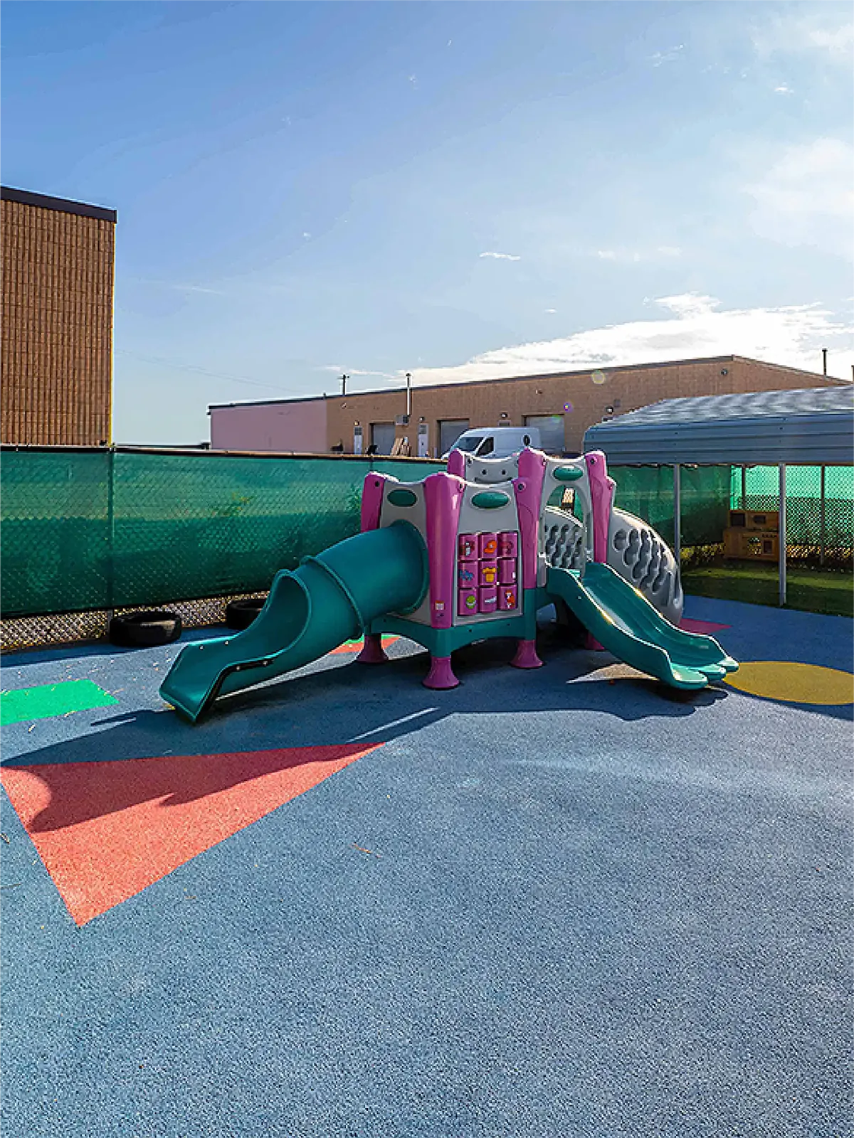 A colorful playground with slides and climbing features stands on a soft rubber surface, surrounded by a green fence—perfect for children at the age for junior kindergarten or families seeking early learning centres.