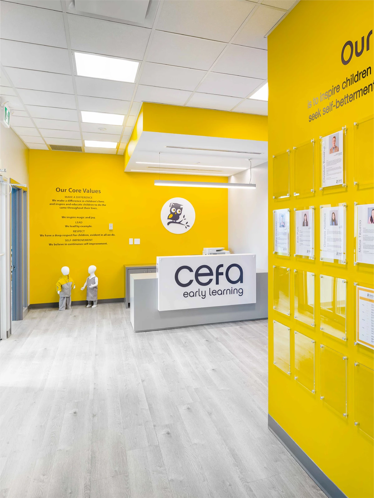 Bright, modern reception area of CEFA Early Learning, one of the leading early learning centres, features yellow walls, a white and gray front desk, staff profiles on display, child mannequins near the entrance, and core values written on the wall.
