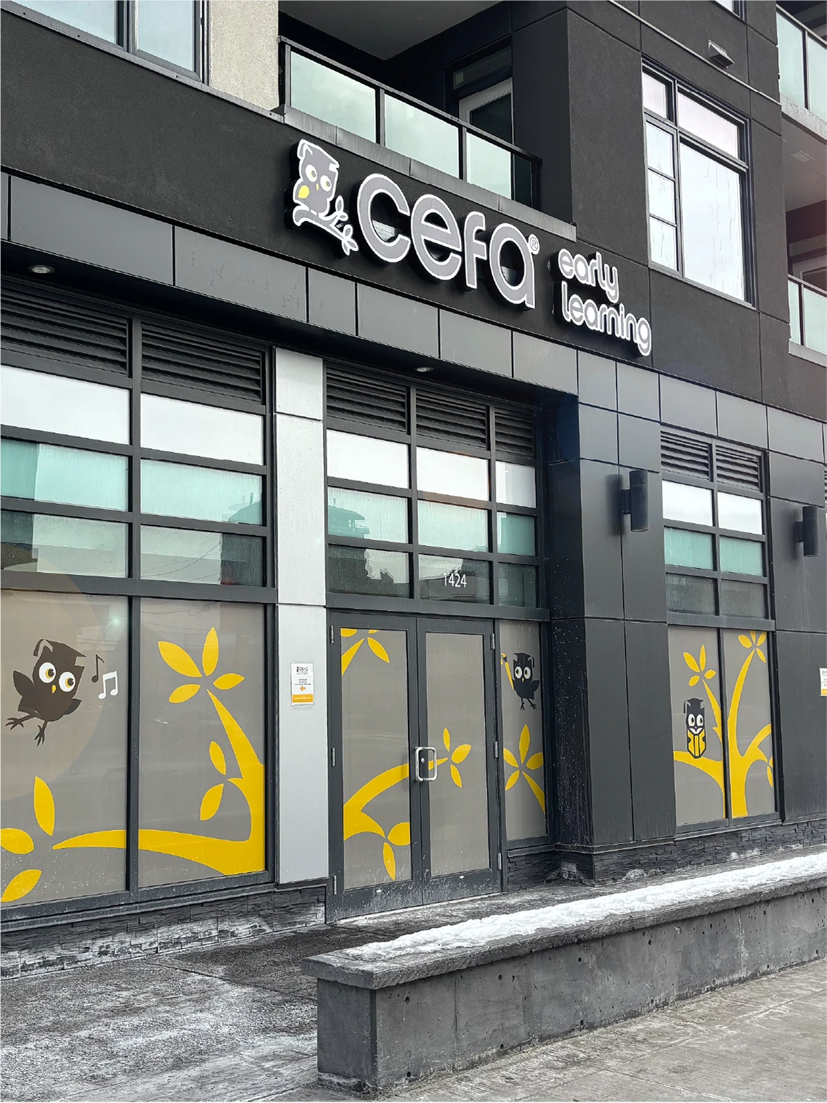 A modern building exterior with large windows displays yellow tree and owl graphics, highlighting one of the top early learning centres. The sign above reads "cefa early learning." A thin layer of snow covers the ground and entrance ledge.