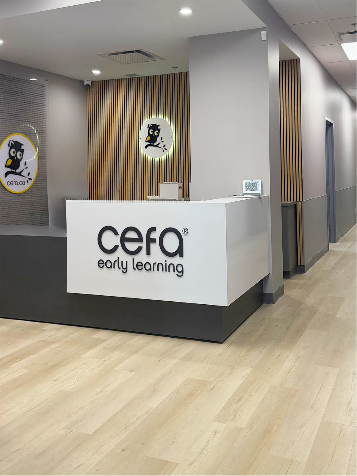 Reception area of a CEFA early learning centre with a modern white desk, wood accents, and owl logos on the wall. The hallway on the right features light wood flooring and gray walls—perfect for families seeking quality preschools close to me.